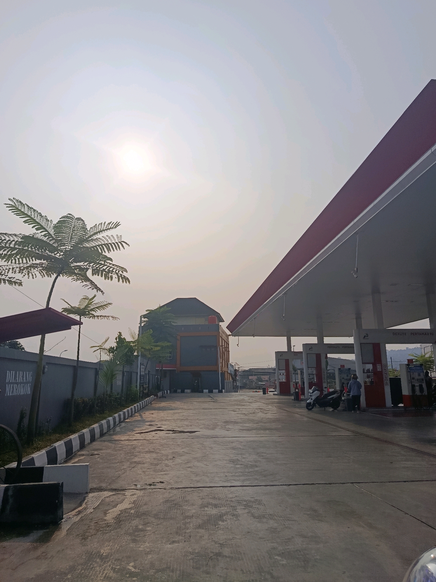 The image depicts a gas station in Samarinda, Indonesia, under a bright, hazy sky. The foreground features a concrete forecourt with distinct expansion joints and a painted curb that alternates between black and white. To the left, a grey concrete wall bears the white text "DILARANG MEROKOK" (No Smoking). Several palm-like trees are visible, including a large one on the left and a smaller one to its right, situated behind a red awning.
The main subject of the scene is the gas station itself. A red-and-white awning covers the fuel pumps, supported by white pillars. Several fuel pumps are visible, with signage indicating fuel types such as "DEXLITE PERTAMAX92". Two people are present at the gas station: one person, wearing a light blue top and grey pants, is standing next to a white scooter, seemingly fueling it. Another person, dressed in dark clothing, is standing near a scooter on the right side of the frame.
Behind the gas station, a multi-story building with dark grey walls and orange accents is visible. Trees and other structures, including what appears to be a bridge in the distance, are also present in the background. The overall environment suggests a typical urban setting in Indonesia during daylight hours, with a slightly overcast or hazy atmosphere.