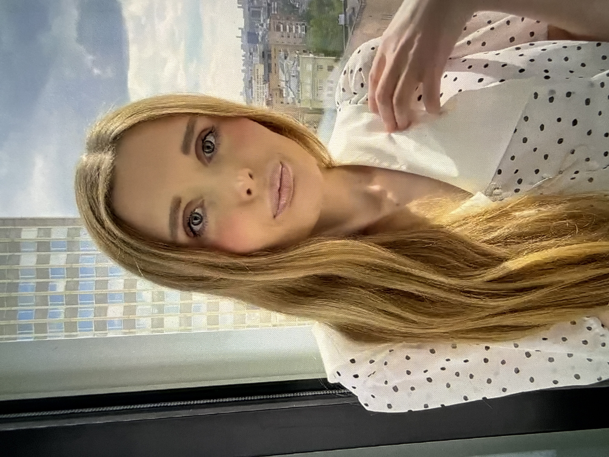A woman with long blonde hair is looking at the camera. She is wearing a white dress with black polka dots. Her face is framed by her hair, and her eyes are looking directly at the viewer. Her lips are slightly parted, and she appears to be speaking or about to speak. Her right hand is raised to her neck, near the collar of her dress. In the background, there is a blurry cityscape with buildings and a cloudy sky. The image is taken from a close-up perspective, focusing on the woman's face and upper body.