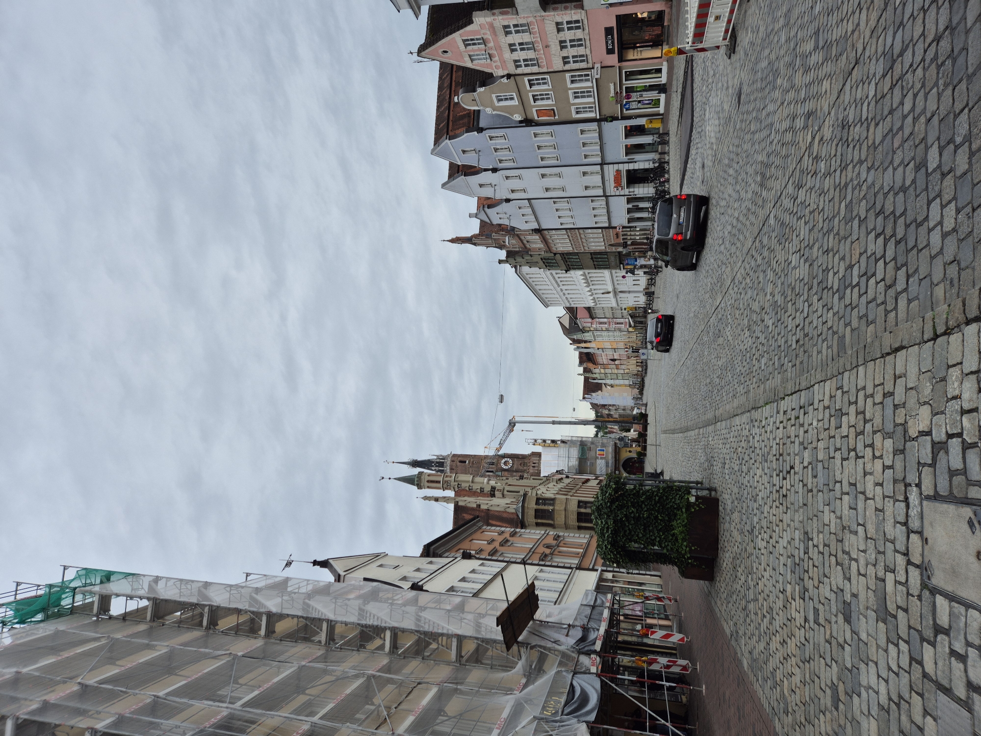The image shows a street scene in Landshut, Germany, likely the Altstadt, given the prominent architecture and cobblestone street. A multi-story building on the left is covered in scaffolding and protective netting, indicating construction or renovation work. The street is paved with cobblestones and has trolley tracks. Several cars are present on the street. On the right side of the street, a row of colorful, traditional German buildings with pitched roofs line the street, some housing shops and businesses. In the distance, a tall church tower with multiple spires and a clock is visible. The sky is overcast and cloudy, suggesting a temperate or cool day. There are no visible people in the image. The overall impression is of a historic European city undergoing some modernization or maintenance while preserving its architectural character.