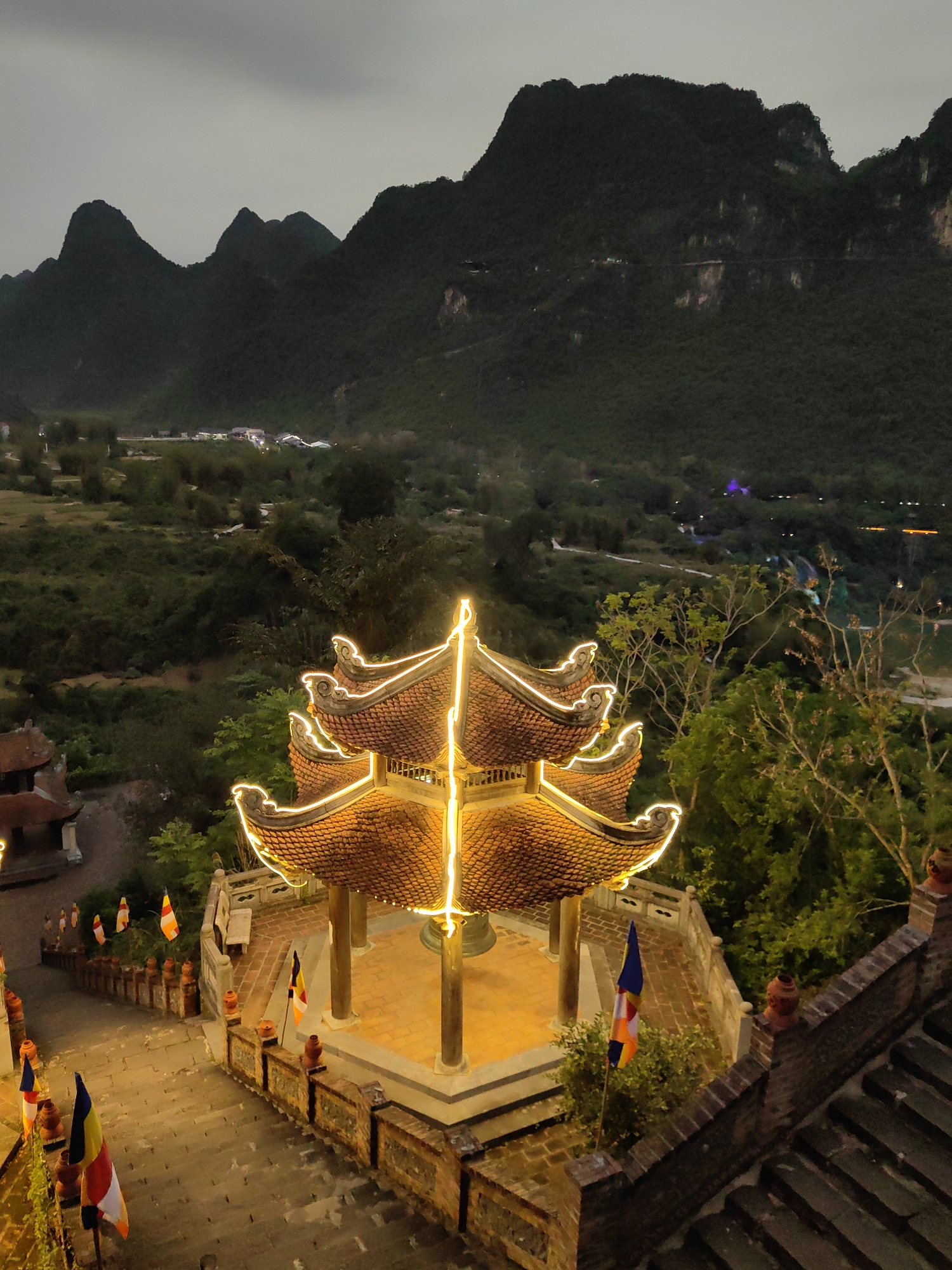 Captured at dusk in Xã Đàm Thủy, Vietnam, the image presents an elevated perspective of a traditional multi-tiered pavilion with curved eaves, illuminated by integrated golden lights. The pavilion is constructed of brick and features ornate detailing. The photograph is taken from an elevated viewpoint looking down onto steps and a courtyard containing the pavilion. The steps and courtyard are lined with Buddhist flags. Behind the pavilion stretches a vista of densely vegetated valleys and distant, mountainous terrain. The overall ambiance is one of serene contemplation amidst a culturally significant landmark.