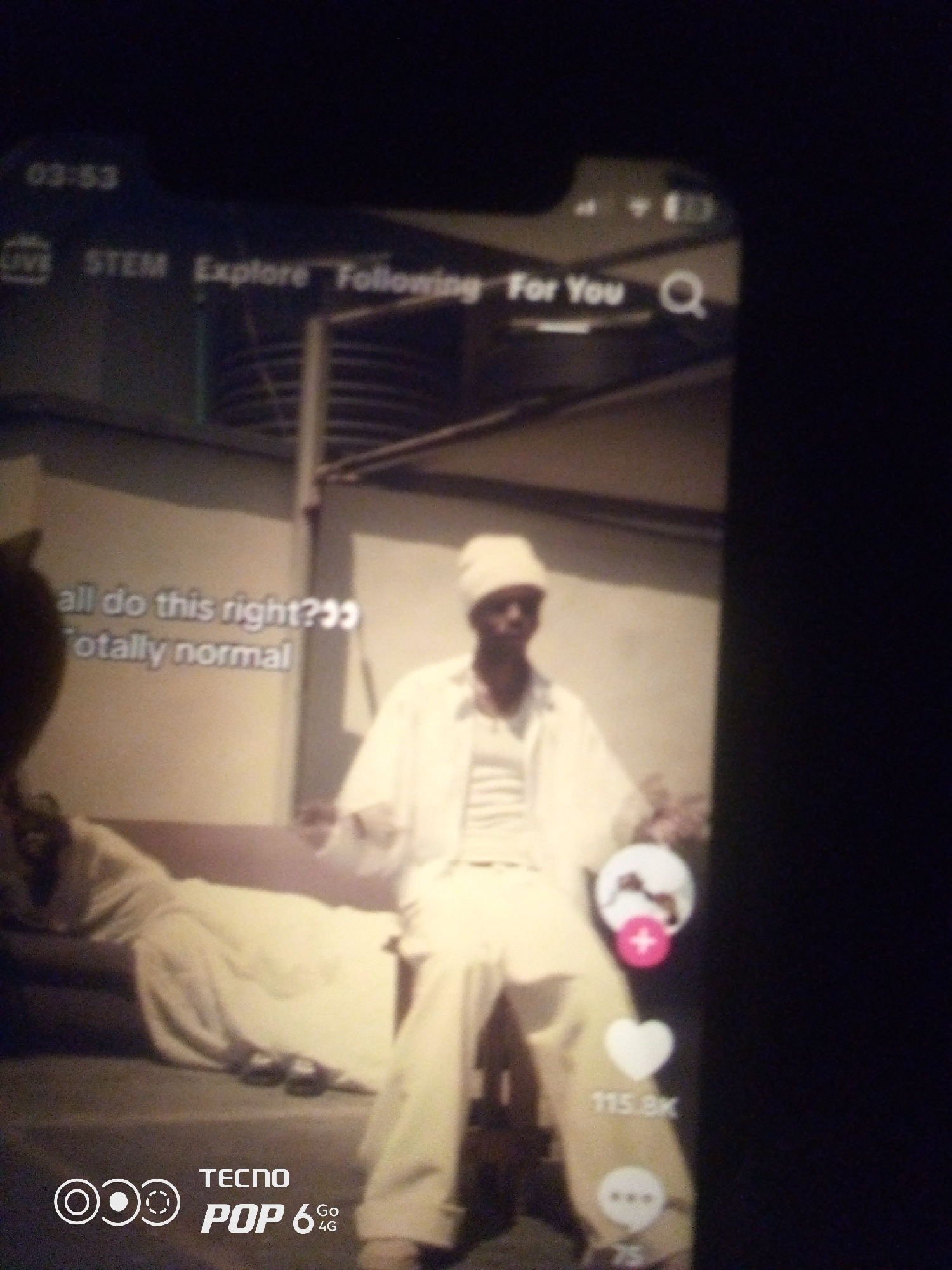 The image is a screenshot of a social media video, likely TikTok, displayed on a mobile phone. The video features a person wearing white clothing and a white head covering, standing and possibly dancing. Text overlays on the video include "all do this right???" and "totally normal". In the top left corner, a timestamp reads "03:53", and there are navigation options like "LIVE", "STEM", "Explore", "Following", and "For You", along with a search icon. On the right side of the video, interactive elements like a heart icon (likely for likes) with the count "115.8K" and a comment icon are visible. The bottom of the screenshot shows a "TECNO POP 6 Go 4G" branding. The background appears to be an outdoor urban setting, possibly a rooftop or an alleyway, with some structures that suggest a city environment. There is no specific indication of the city of Howayi, Nigeria within the image itself.
