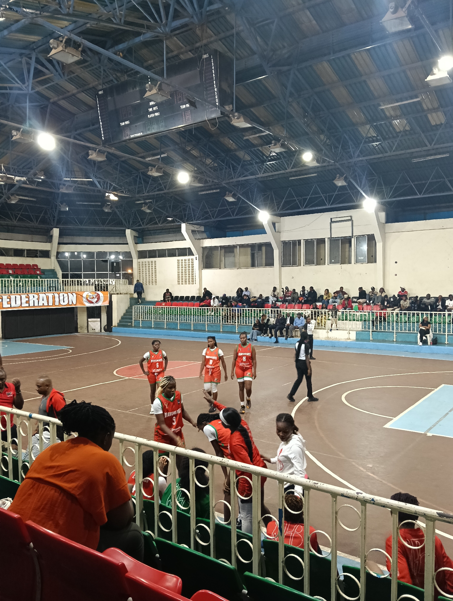 This media file depicts an indoor women's basketball game or practice session taking place in a gymnasium in Nairobi, Kenya. The scene is illuminated by numerous bright overhead lights, suggesting an evening or night event.
**Subjects:** The primary subjects are several female basketball players on the court, dressed in matching red-orange and white uniforms. Some players are identifiable by numbers (e.g., 6, 9, 16) and team names like "BRAVEHEARTS" and "Imports" on their jerseys. A referee in a black and white uniform is also present on the court. In the background and foreground, a diverse group of spectators is seated in tiered stands, watching the activity.
**Setting and Objects:** The venue features a basketball court with distinct brown and light blue sections, marked with white lines. Elevated red and green seating areas are visible, some of which are occupied by spectators. A prominent scoreboard hangs from the ceiling, displaying game statistics. The ceiling itself is composed of metal trusses and corrugated panels, fitted with numerous high-intensity lights. A metal railing separates the front row of spectators from the playing area. A large banner with the text "FEDERATION" and an emblem is visible on a wall beside the court.
**Activity:** The players appear to be in a relaxed state, walking or standing on the court, suggesting a break in play, warm-up, or cool-down period rather than active gameplay. Spectators are observing the scene.
**Notable Details:** The artificial lighting creates strong reflections on the polished court surface. The atmosphere is that of a local sporting event.
**Visible Text:**
* On the scoreboard: "FOULS", "TIME OUTS", "PLAYER-FOULS", "TEAM-FOULS", and the manufacturer's name "DAKTRONICS". Numbers for fouls range from 1 to at least 15.
* On a banner: "FEDERATION".
* On player jerseys: "BRAVEHEARTS", "Imports", and player numbers "6", "9", "16".