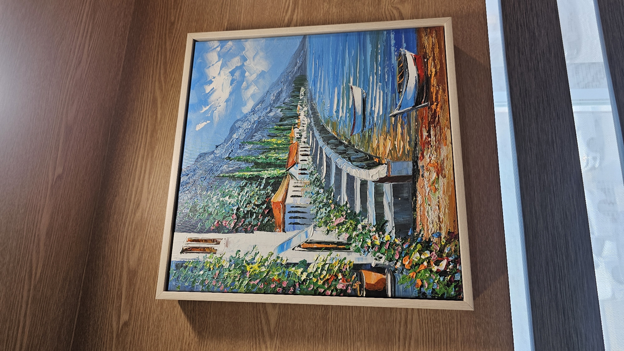 The image displays a framed oil painting of a scenic coastal village. The painting depicts a row of white buildings with orange-tiled roofs lining a cobblestone pathway. Lush green vegetation and colorful flowers cascade down the walls of the buildings and the surrounding steps. A body of bright blue water, likely a bay or lake, stretches out in front of the village, with two small boats visible on its surface. In the background, a majestic, mist-covered mountain rises against a sky filled with dynamic white clouds. The painting is displayed on a wooden wall, with a portion of a window blind visible at the bottom. There is no indication that this scene is located in Asan-si, South Korea.
