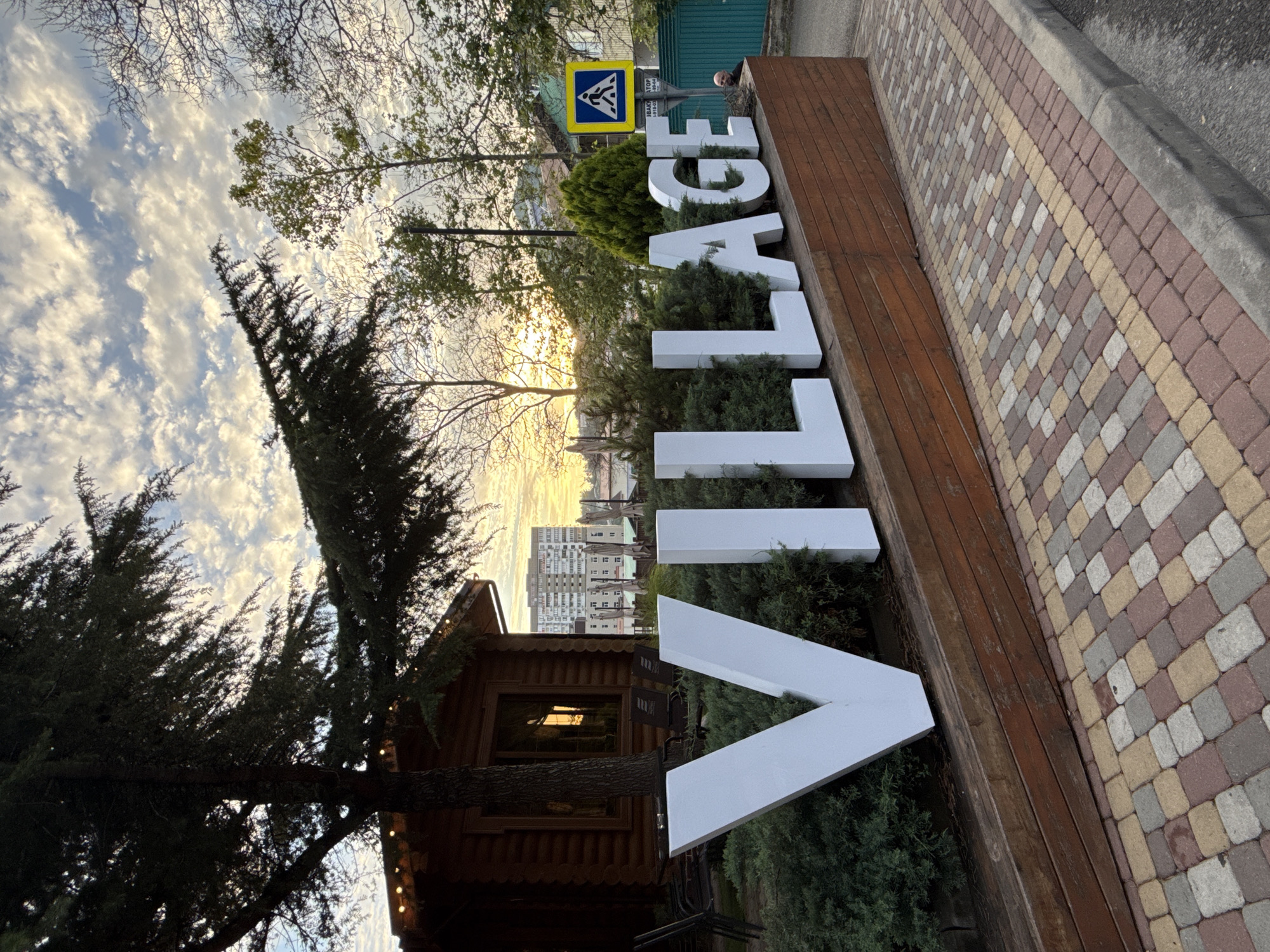 A wide-angle, eye-level outdoor shot captures the large white letters spelling out "VILLAGE" in front of a wooden structure. The letters are placed in front of manicured green bushes and on a sloped wooden planter box. Behind the letters, a rustic wooden building with a lit string of lights along its roofline is visible, with chairs and tables outside. Further in the background, a tall residential building with many balconies is seen. The sky above is filled with a pattern of white clouds against a pale blue sky, with a hint of sunset orange peeking through on the right. The ground in the foreground is paved with colorful interlocking bricks. A pedestrian crossing sign is visible on the right side of the image, near the residential building. A man's head and shoulders are visible in the far right background, looking to the left. The scene is likely in Olginka, Russia, given the context provided.
