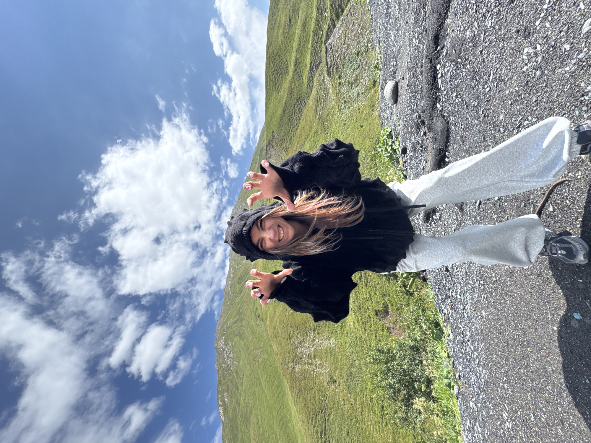 A young woman is pictured in a playful pose with her hands raised in front of her, mimicking claws, against a backdrop of lush green mountains under a blue sky dotted with white clouds. She is wearing a black hooded jacket with textured sleeves, light gray sweatpants, and athletic shoes. Her hair is blonde with pink streaks. The ground beneath her is covered in dark gravel and small rocks. The environment suggests a mountainous, possibly rural, area. The image does not provide any specific details to indicate it is located in the city of Almasiani, Georgia.