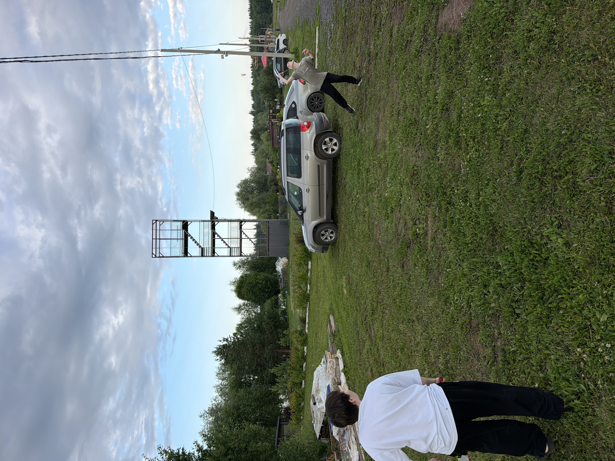 This image, taken in Bol'shaya Ven'ya, Russia, depicts an outdoor scene on a cloudy day, likely in the late afternoon or early evening given the lighting. The foreground features a person from behind, wearing a white t-shirt and black pants, standing on a grassy area. Further back, another person, dressed in a green t-shirt and black pants, is captured mid-air in a dynamic pose, as if jumping or falling, with their arms outstretched. This person is positioned near a silver SUV and a dark-colored car.
In the background, a multi-story metal structure with stairs is visible. The setting appears to be a rural or semi-rural area, with abundant green grass, trees, and some scattered wooden structures or debris. The sky is filled with a mix of grey and lighter clouds, suggesting a changeable weather pattern. The overall impression is one of an active moment, possibly a recreational activity or a playful stunt taking place in a somewhat informal outdoor setting. There is no visible text in the image.