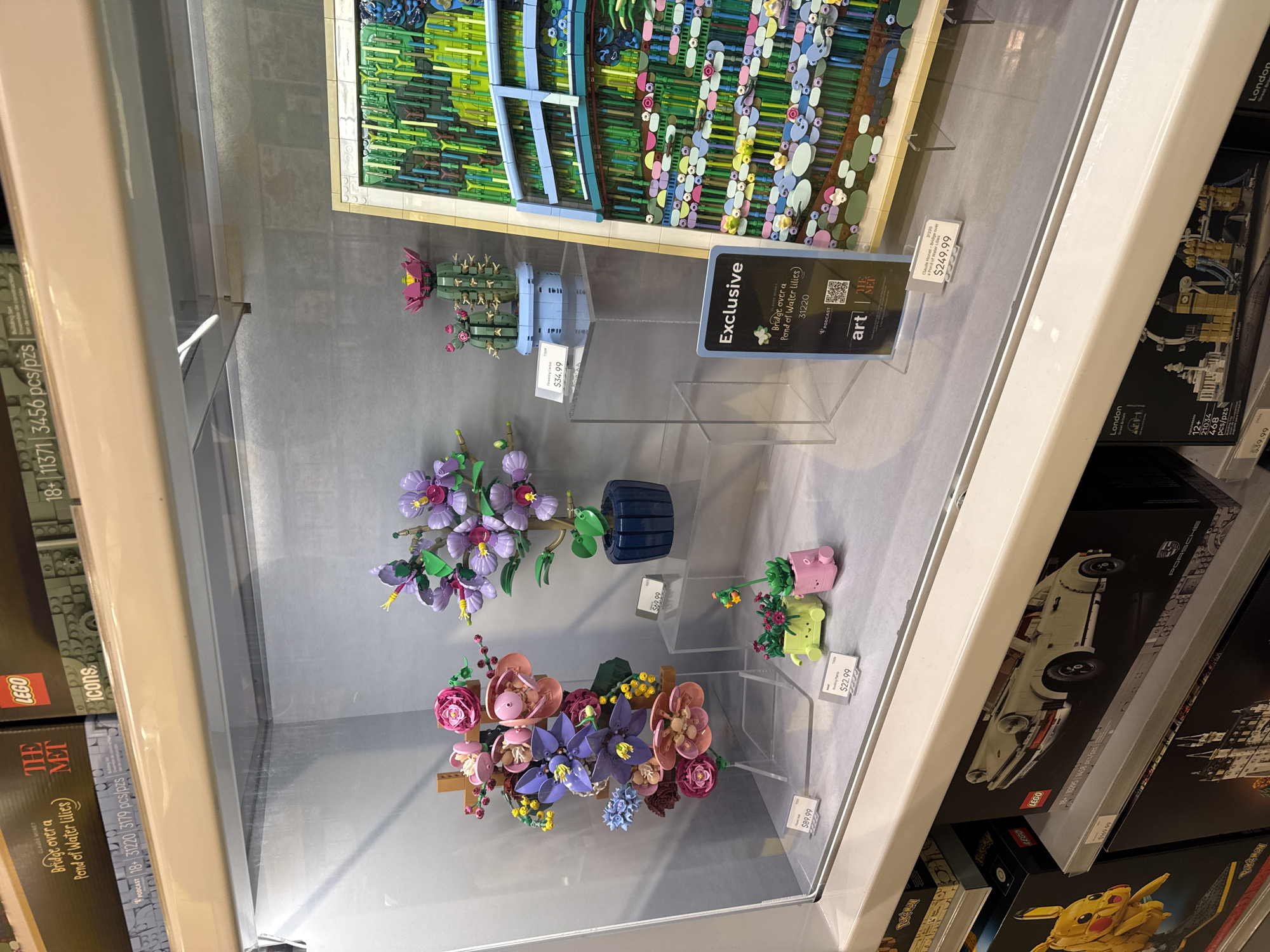 This image depicts a display case containing various Lego sets and floral Lego creations. The location is identified as Schaumburg, United States of America, based on the presence of products sold within a general retail environment.
Within the display case, there are three floral Lego arrangements and a Lego cactus, each on a separate clear stand. The arrangements consist of various colorful Lego bricks to mimic real flowers. Price tags are visible near each arrangement, indicating the retail price. Above the Lego flowers, the display contains Lego sets for sale including Lego Art's rendition of Claude Monet's "Bridge over a Pond of Water Lilies," priced at $249.99, and other Lego sets, including a Lego Great Britain-themed set of the London skyline, priced at $39.99. The box next to it features a Lego Porsche.
The display case is positioned next to shelves stocked with various other Lego sets. A sign that identifies the Lego store's exclusive Lego Art collaboration with the Metropolitan Museum of Art is visible, which further implies the setting is a retail store selling Lego products.
