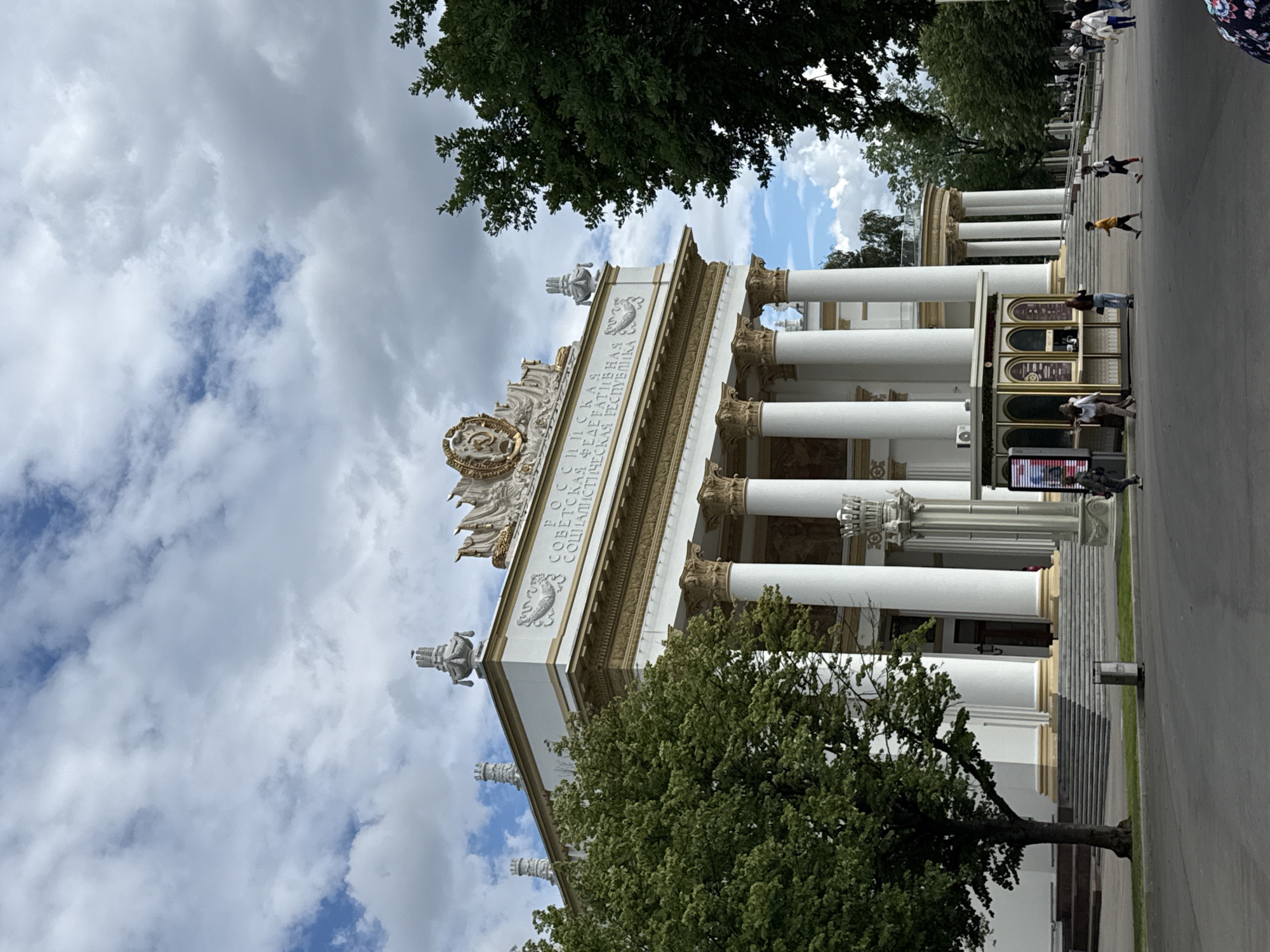 The image shows an outdoor scene featuring a large, ornate white building with classical architectural elements, including columns and decorative reliefs. Green trees are visible in the foreground and background, and the sky is partly cloudy with patches of blue. Several people are present, walking along a paved pathway next to the building and a set of stairs. One person is leaning against a kiosk-like structure. The building appears to be a pavilion or exhibition hall.
The text "РОССИЯ СОВЕТСКАЯ СОЦИАЛИСТИЧЕСКАЯ РЕСПУБЛИКА" is visible on the facade of the building, indicating it is likely from the Soviet era and located in Russia. The scene suggests a daytime setting, possibly a warm or mild day given the presence of trees and people in casual attire. The overall atmosphere appears to be that of a public park or exhibition ground, with visitors enjoying the surroundings. The orientation of the image is portrait, with the scene appearing to be tilted to the right.