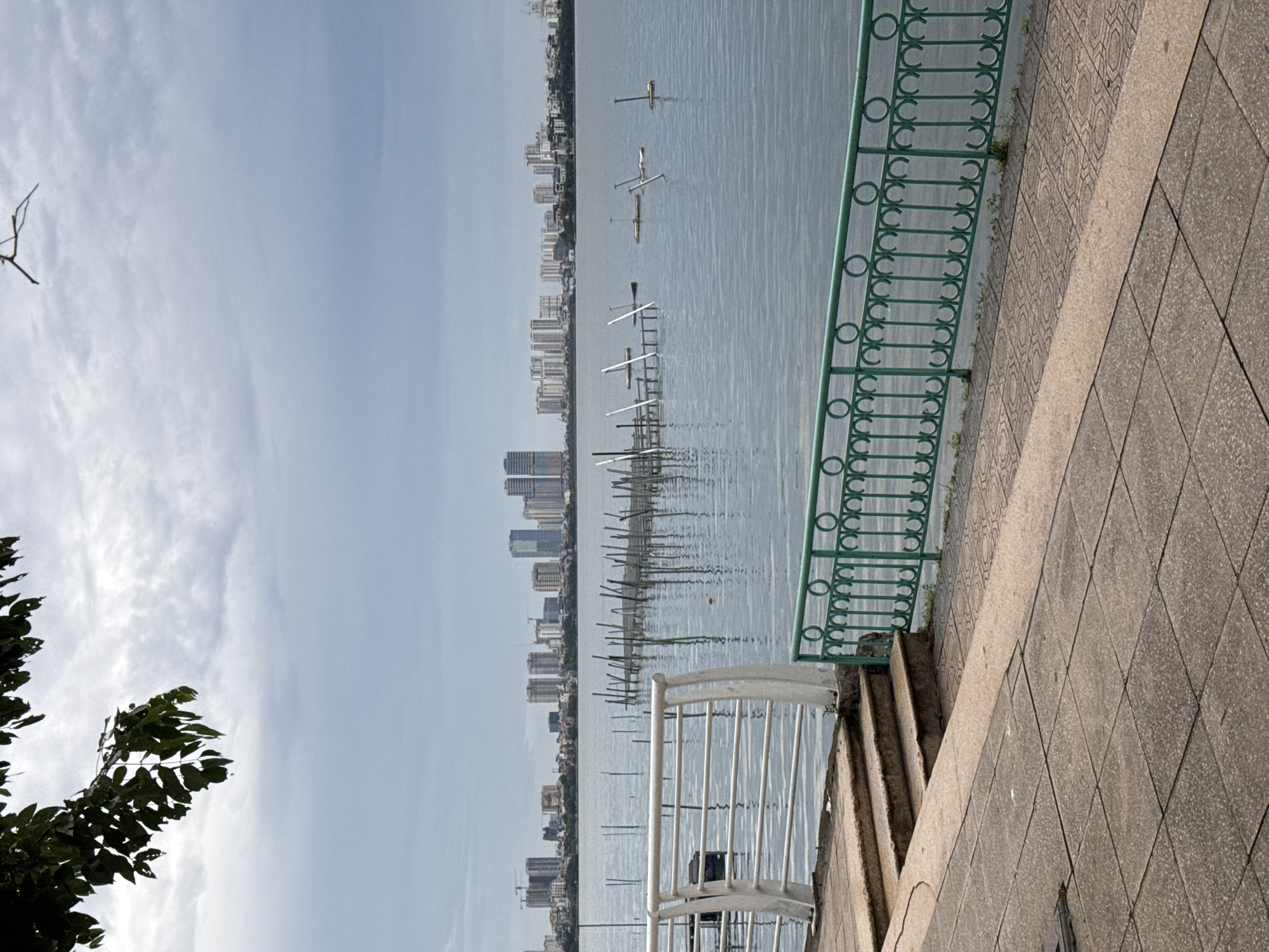 The image depicts a scene along West Lake in Hanoi, Vietnam, on an overcast day. In the foreground, a paved walkway with large square tiles transitions to a decorative tiled area. A set of concrete steps leads down to a path with a white metal railing. Adjacent to this is a green metal fence with circular and vertical decorative elements, bordering a body of water.
The water of West Lake stretches across the middle ground, with several wooden poles and structures visible, some appearing to be part of a fishing or aquaculture setup, and others resembling simple posts sticking out of the water. In the background, the Hanoi skyline is visible, characterized by a dense arrangement of modern high-rise buildings of varying heights and architectural styles, suggesting an urban waterfront. The sky is cloudy and grey, creating a subdued atmosphere. A few tree branches with dark green leaves overhang the top left corner of the frame.