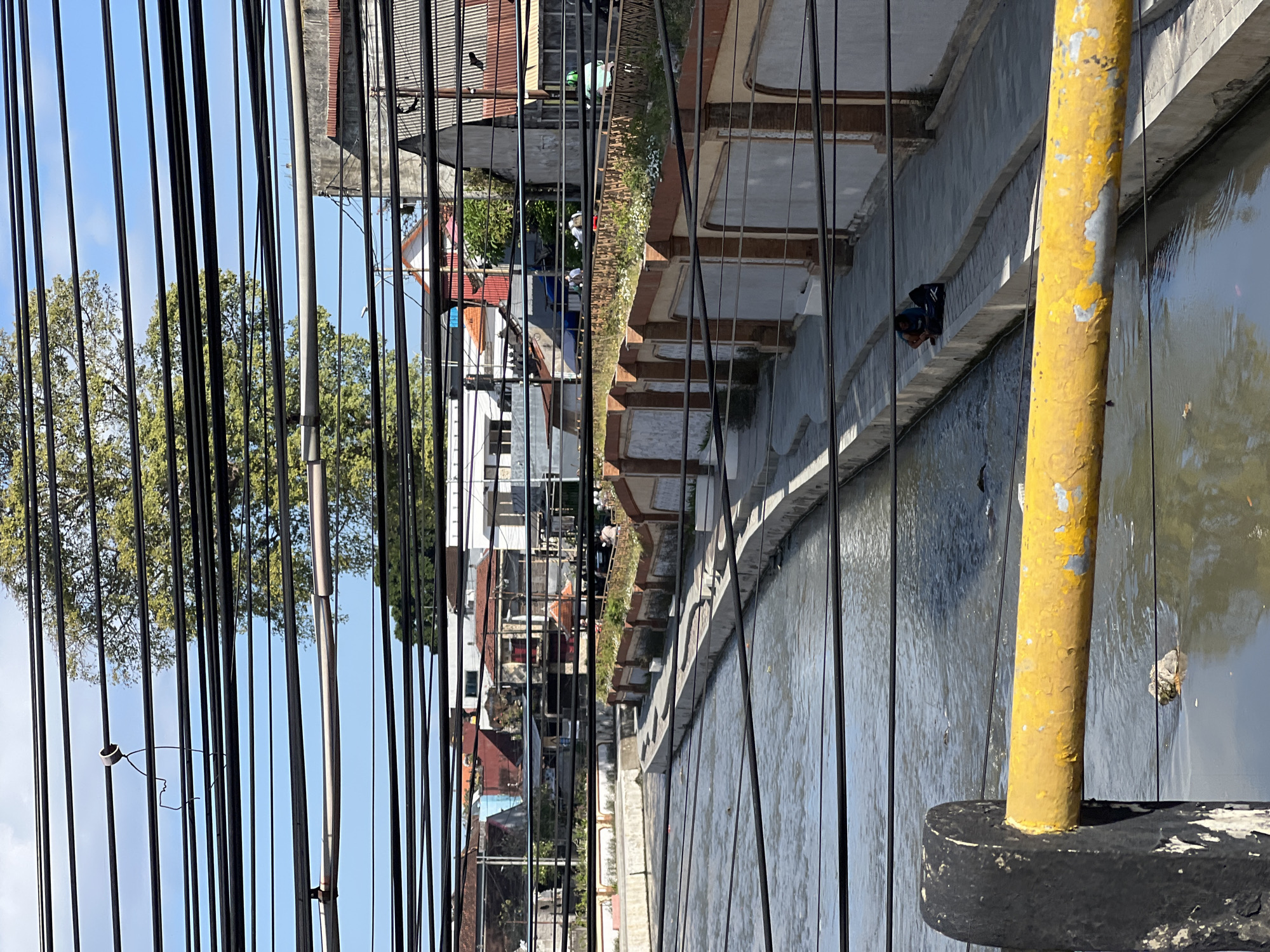 The image depicts a view of a canal in Denpasar, Indonesia, from a low vantage point, partially obscured by numerous overhead electrical wires. The canal is lined with concrete pathways and decorative walls with arched patterns. In the middle distance, two individuals are seated on the canal's edge, one with a dark backpack. The canal water is murky and contains debris. Beyond the canal, buildings with red tile roofs and white facades are visible, interspersed with lush green trees. The sky is a clear blue with some clouds. In the foreground, a yellow-painted metal railing with black supports is prominently displayed, further framing the scene.