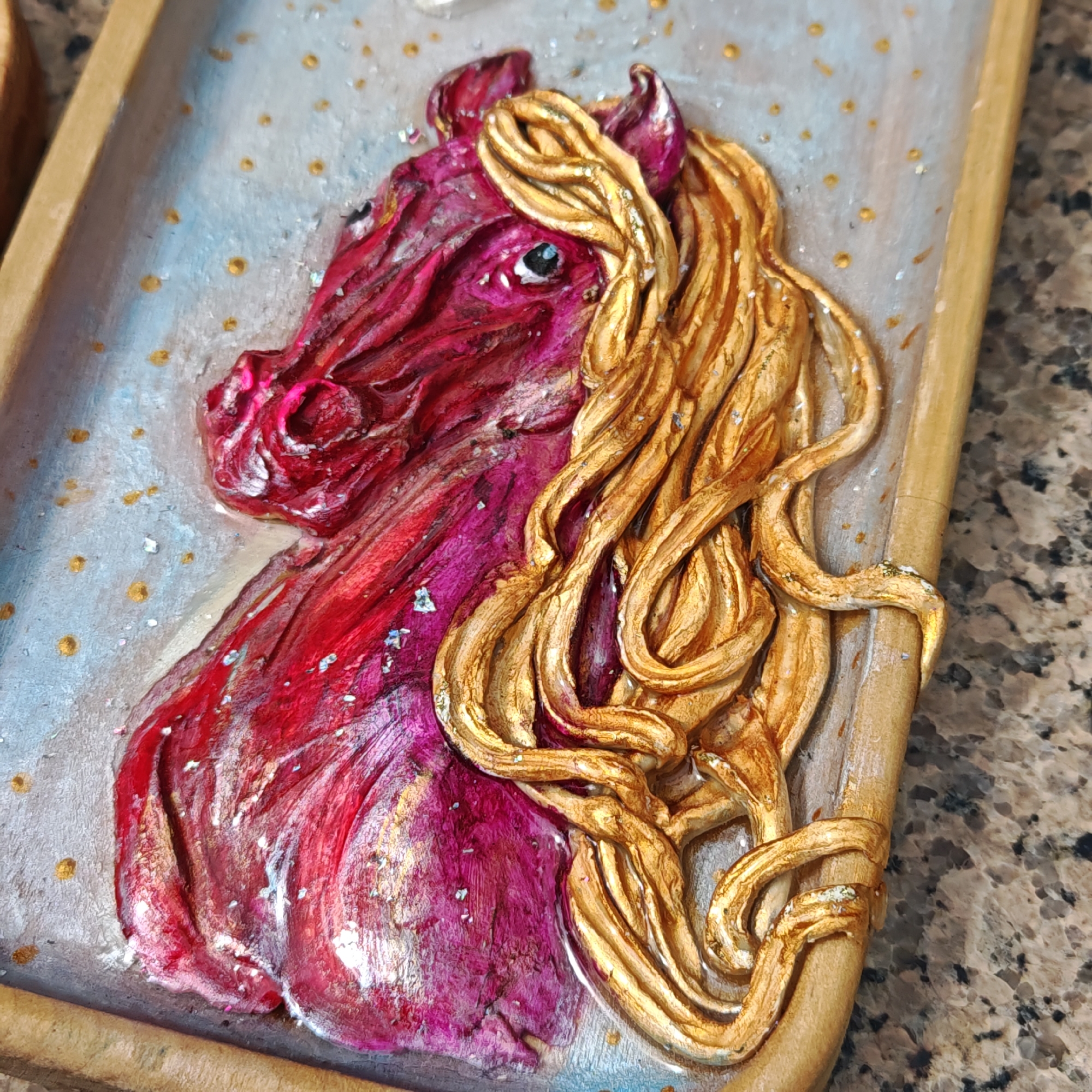 A close-up shot depicts a three-dimensional artwork featuring a horse's head, predominantly red and gold in color, set within a wooden frame. The horse's mane is styled in flowing, sculpted strands, painted gold, contrasting with the red and burgundy hues of the horse's head and neck. Metallic flecks are visible on the horse and the surrounding background. The background is a light gradient of blue and white with small gold dots scattered across it. The frame is simple, made of light-colored wood, and the entire piece rests on a mottled brown surface. This artwork is not verifiably located in Taganrog, Russia, and there is no evidence of people or actions within the scene.