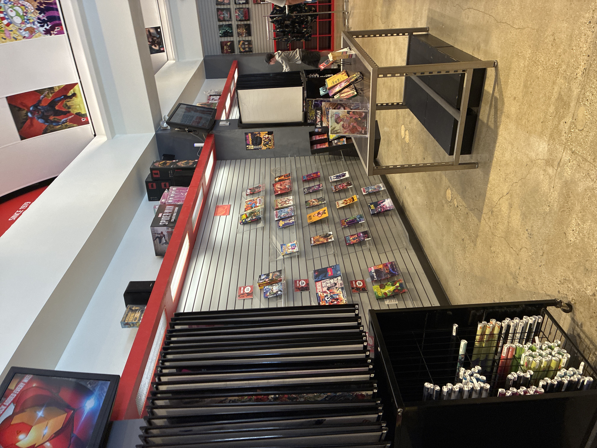 The image depicts the interior of a comic book store. A wall-mounted display of comic books is visible, organized on horizontal shelves against a grey, slatted background. Several comic books are displayed in clear plastic sleeves. To the left, a tall, black rack is filled with what appear to be rolled-up posters or comic book precursors, each individually wrapped and tagged. Above the comic book shelves, a red shelf displays various comic book boxes, including one labeled "SPIDER-MAN." A television screen is mounted on the wall, showing an image of Iron Man. The ceiling features white panels and some red accents. A metal display table with black shelves is positioned in the foreground, holding a few comic books and other merchandise. A young person wearing a grey hoodie and a black baseball cap is standing near the display table, facing away from the camera. In the background, more comic book artwork is displayed on the walls, and a rack of clothing is visible. The floor is a polished concrete surface. The scene is brightly lit.