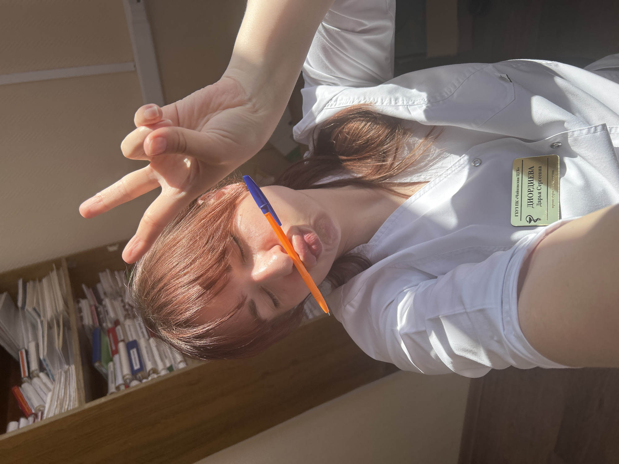 A woman in a white medical coat poses with a pen held between her lips, while making a peace sign with her hand. The woman has red hair and a name badge on her coat, which reads “ГУЗ ПК «Чайковская ЦГБ» ДИОРДИЕВА Дарья Сергеевна”. There is a shelf of medical supplies visible in the background. While the image description says the woman is located in Фоки, Russia, the name badge indicates she works at the "Chaikovsky Central City Hospital".