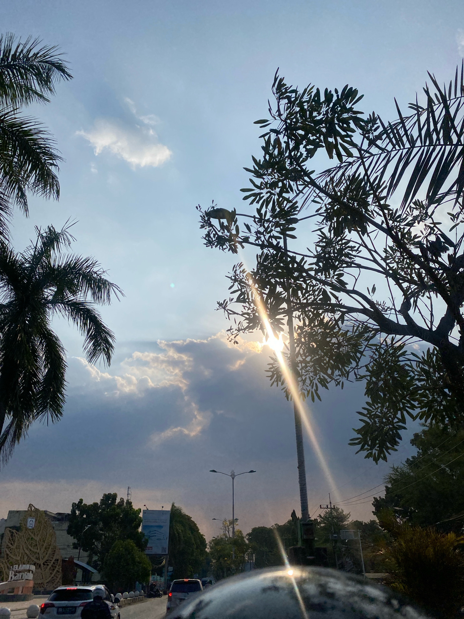 The image depicts a street scene in Banjarbaru, Indonesia, under a sky with scattered clouds and visible sun rays. Palm trees are present on the left, and a tree with broad leaves is on the right, both silhouetted against the sky. In the background, a street with several vehicles, including cars and motorcycles, is visible. Buildings and vegetation line the street. A large woven structure with text that appears to say "SELAMAT BANJAR..." is on the left side of the road. The sun is partially obscured by clouds, casting strong light rays through the scene. The overall impression is that of an urban environment during daylight, possibly late afternoon or early morning due to the angle of the sun.
