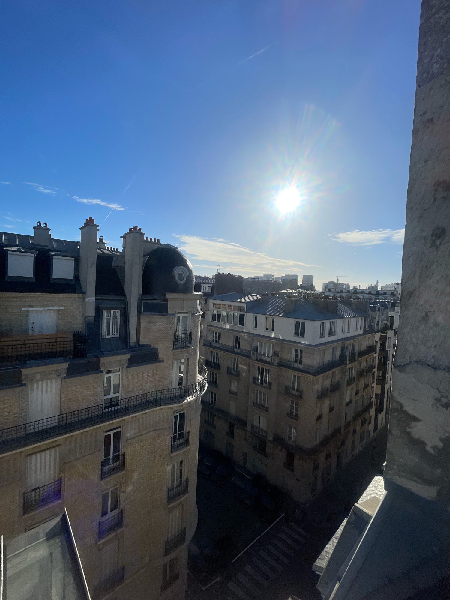 The image captures a daytime view of Parisian rooftops and buildings under a bright, sunny sky with scattered clouds. The sun is a prominent feature, high in the sky and casting a strong glare. In the foreground, to the right, a large stone architectural element, possibly part of a building or a chimney, partially obscures the view.
The middle ground features several apartment buildings with characteristic Parisian architecture. These buildings are characterized by light-colored brickwork, dark mansard roofs with dormer windows and chimneys, and regularly spaced windows, many with balconies. One building in the left-middle ground has a distinctive curved facade and a domed section on its roof. Another building, further back and to the right, appears to have a similar architectural style. The perspective suggests the photo was taken from a higher vantage point, looking down and across the cityscape.
In the background, a denser urban landscape with taller buildings is visible, along with a construction crane, indicating ongoing development. The street level below is visible in some sections, showing a crosswalk and the dark shapes of parked cars, all cast in shadow by the overhead buildings and the low angle of the sun. A faint contrail from an aircraft is visible in the sky. The overall impression is of a typical urban scene in Paris on a clear, sunny day.