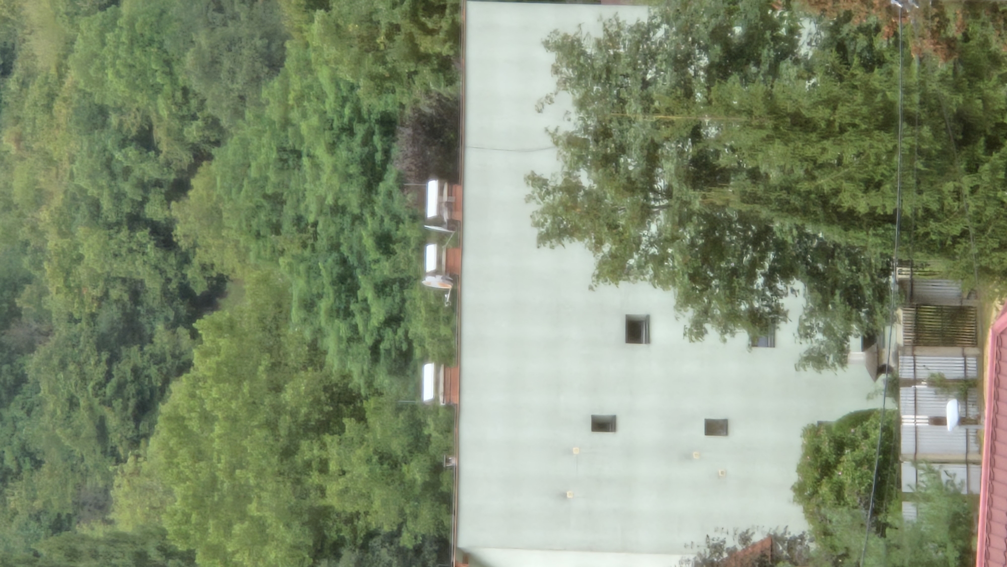 The image displays a section of a building facade with a pale green color. Three rectangular windows are visible on the facade. On the roof of the building, three chimney stacks are present, two of which are topped with white cylindrical structures. Above the building, a dense green forest covers a hillside, with some trees appearing lighter green than others. In the foreground, a portion of a fence is visible, constructed from vertical metal bars and white panels. To the left of the fence, a portion of a red roof is visible. The scene appears to be an exterior view in a built-up area adjacent to a forested hillside, likely in the city of Prešov, Slovakia, given the building style and surrounding vegetation.