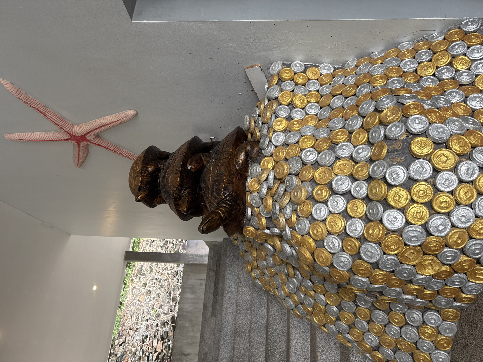 The image shows a statue of three turtles stacked on top of each other with a covering of silver and gold-colored coins, located in ตำบลเกาะพะงัน, Thailand. The statue is positioned inside a building, next to a stairway leading outside where there are plants and rocks. A starfish sculpture is attached to the wall near the turtles.