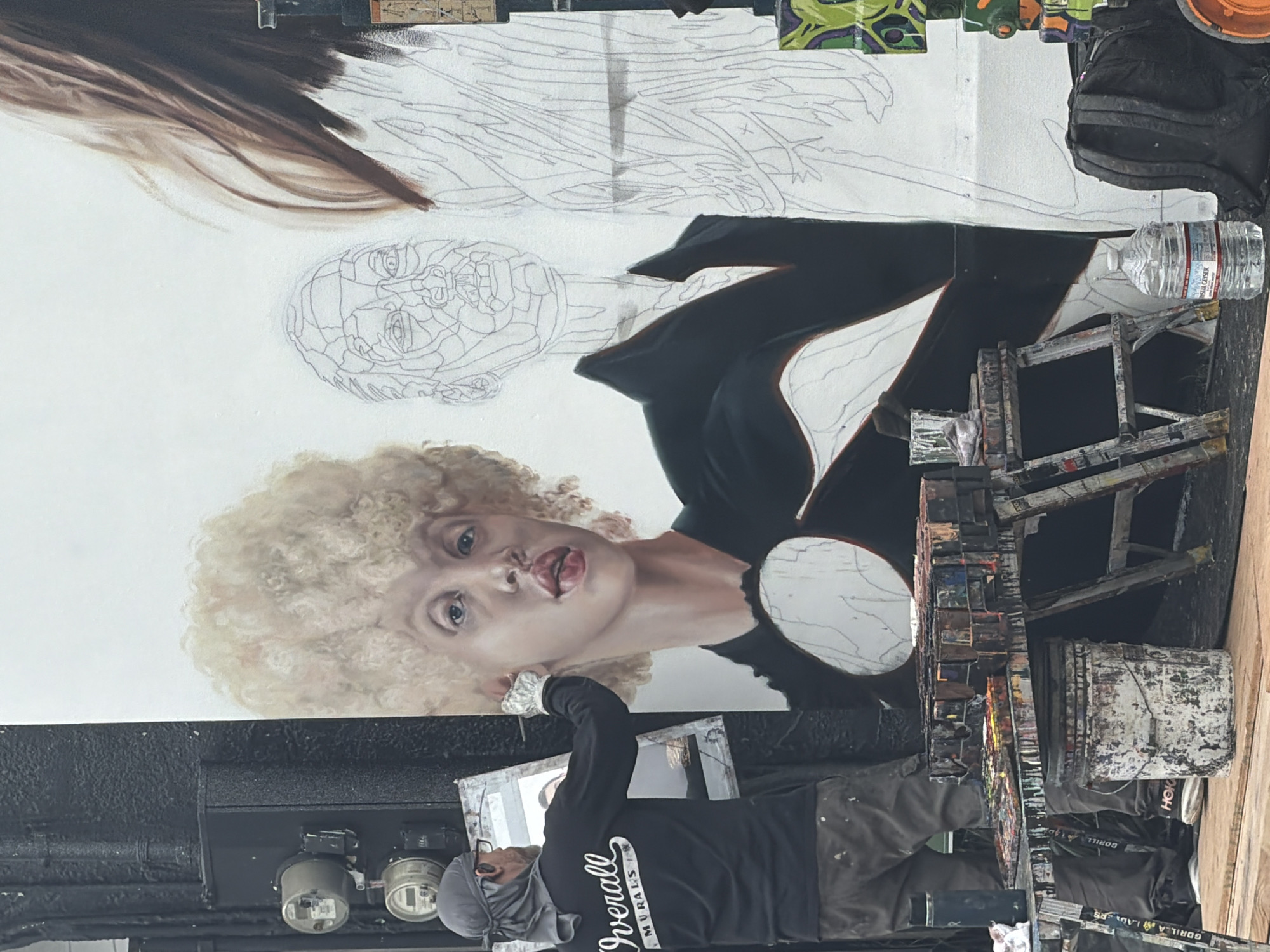 A mural of a woman with blonde curly hair and fair skin is being painted on a building in Los Angeles, United States of America. The mural is only partially complete, with the face of the woman filled in with color, a line drawing of a second face above, and a line drawing of the rest of the body above that. The woman in the mural is wearing a black jacket. An artist wearing a grey head covering, glasses, a black shirt with the words "Overall Murals" on it, and grey pants is standing in front of the mural and working on it. A partially painted painting on a canvas is propped up in front of the artist. Various painting supplies, including paint cans, palettes, and scaffolding, are also visible in the foreground. Electrical boxes can be seen on the side of the building near the artist.