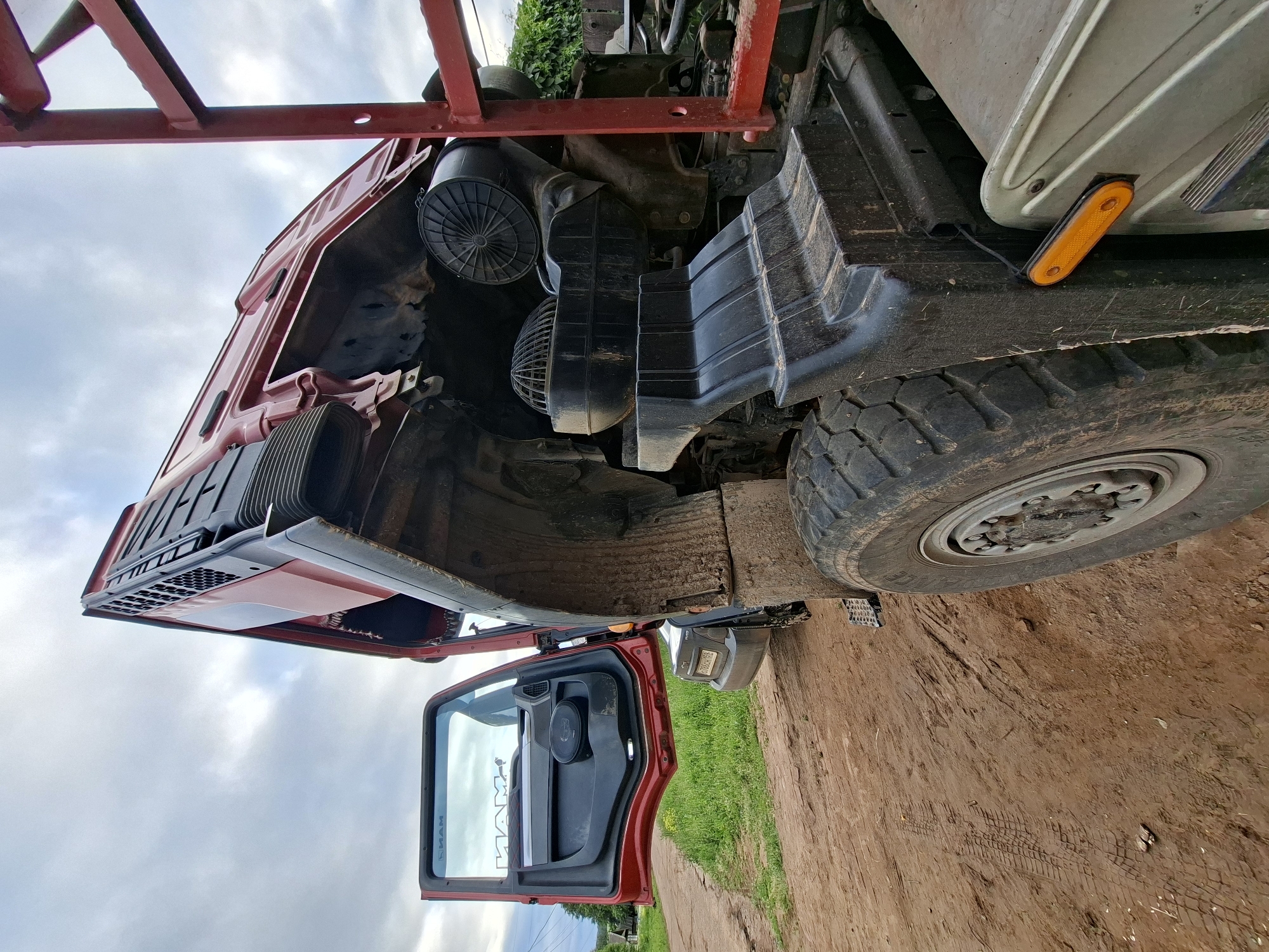 A red MAN heavy-duty truck is depicted on a reddish-brown dirt road with its cab tilted forward, exposing the engine bay. The driver's side door is wide open, revealing a black interior panel with a speaker and a reflected "MAN" logo on the window glass. The truck's front right tire, deeply treaded and covered in dirt, is prominent in the foreground. Adjacent to the tire, a black muddy fender is visible, along with a rectangular orange side marker light. Within the exposed engine compartment, various black plastic air intake components and a dark engine block are discernible. To the right, a vertical red ladder-like framework, part of the truck's specialized body (possibly for a flatbed or logging), stands against dense green foliage. Behind the open truck door, a dark-colored passenger vehicle with a visible Russian license plate (O 706 CM 152) is partially parked on the dirt track. The immediate surroundings consist of green grass and weeds alongside the unpaved road, which displays tire tracks. The background features more distant green trees, faint power lines, and what appear to be rural structures under a cloudy, overcast sky. No individuals are present in the image. The scene is located in an unspecified rural or industrial area in Unknown, Unknown.