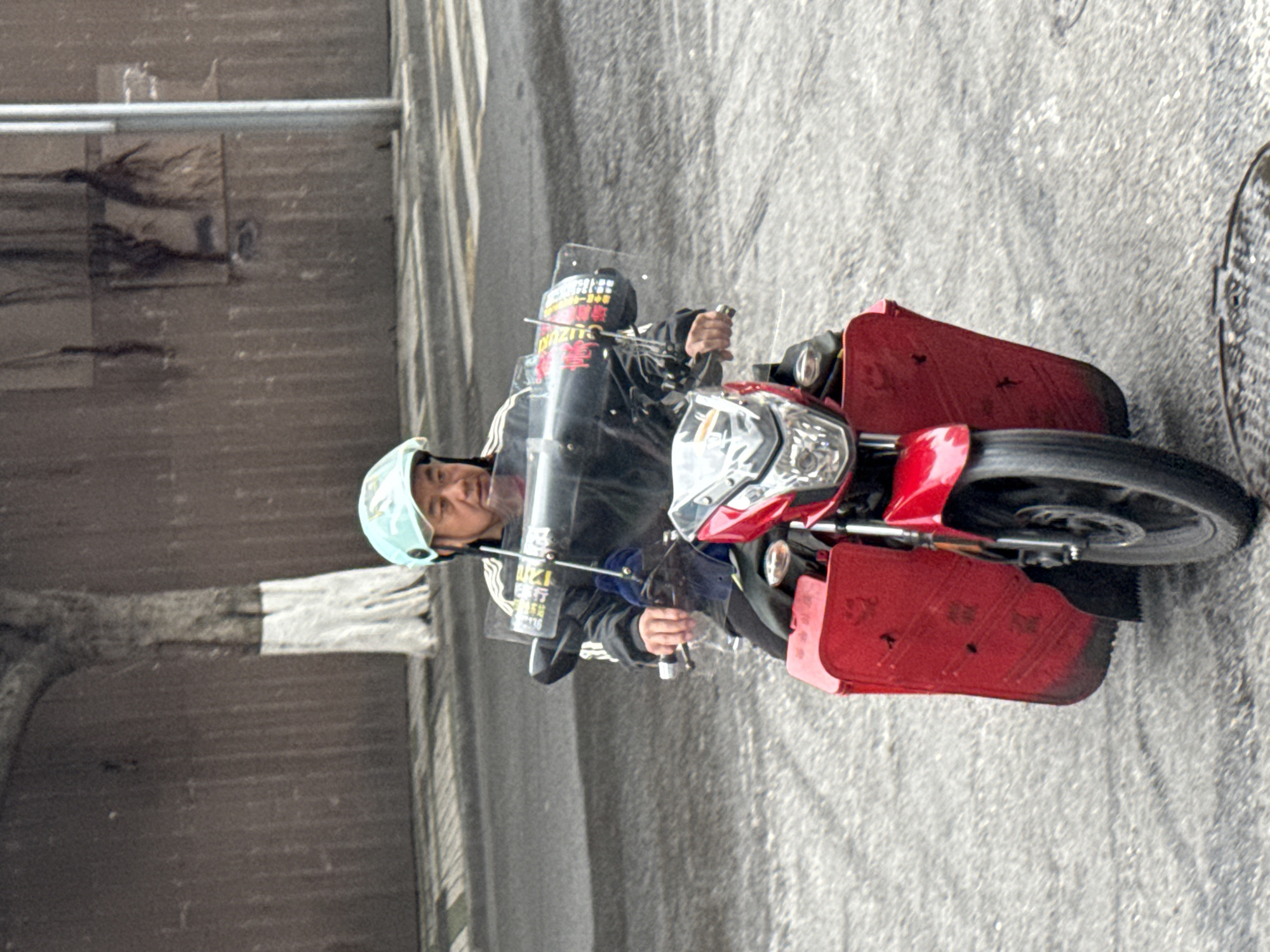 A man wearing a light blue helmet and a black jacket with white stripes rides a red scooter equipped with side panniers on a paved road in the Yuzhong District of China. The background includes a grey concrete wall and a crosswalk. A metal sewer grate is visible on the right side of the frame.