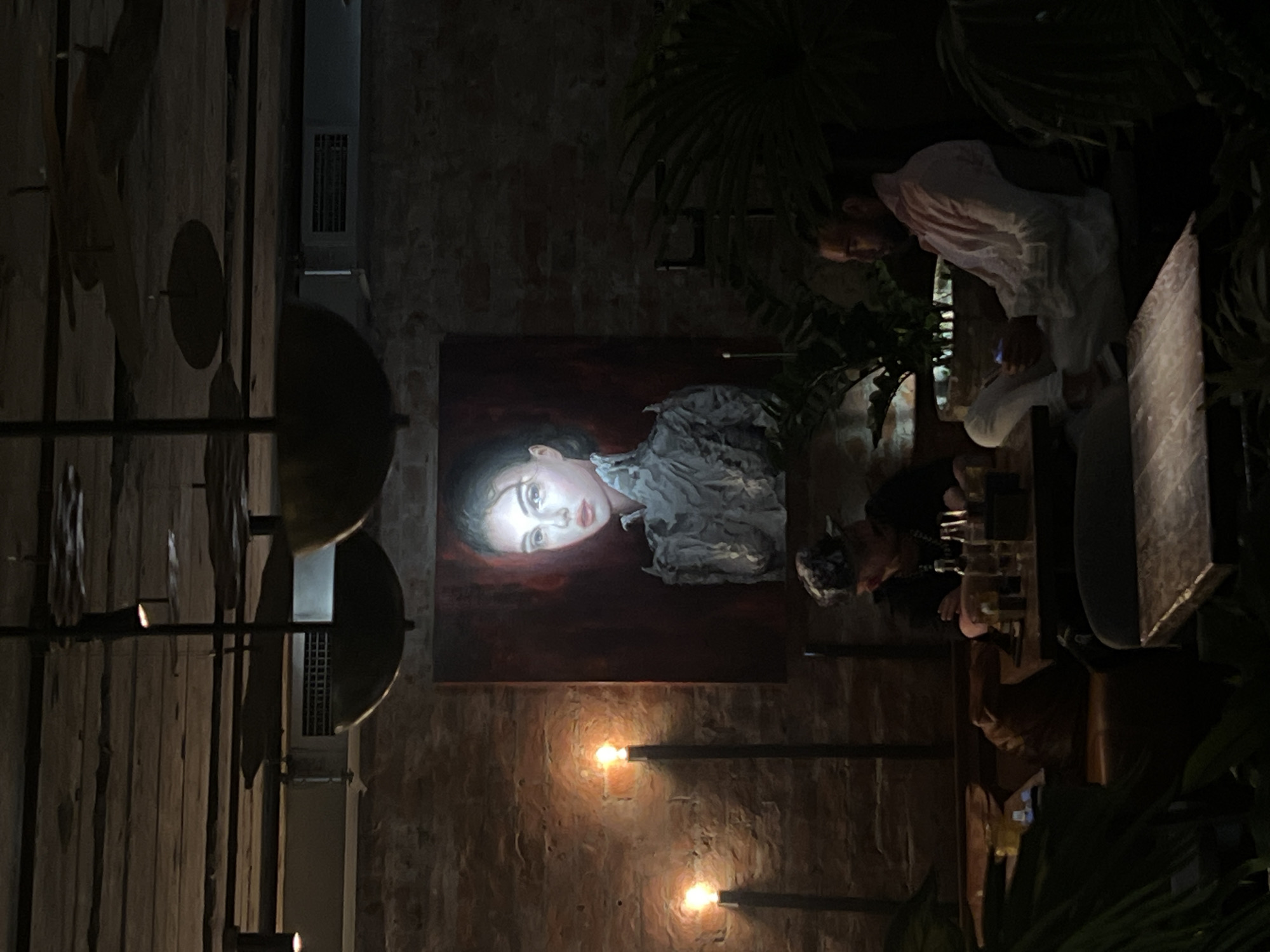 This image portrays the dimly lit interior of a restaurant or bar in Moscow, Russia. The setting features exposed red brick walls and a dark, rustic wooden ceiling with visible beams and planks. Ambient lighting is provided by several warm-toned, exposed filament-style bulbs, including two wall-mounted fixtures on the left and integrated spotlights within large, dark, bowl-shaped pendant lights suspended from the ceiling. A rectangular air vent is visible on the upper central wall. Dark green, leafy plants are interspersed throughout the space, particularly in the foreground and mid-ground.
A large, vertically oriented portrait painting dominates the central brick wall. It depicts a young woman with light skin, dark hair, and a serious expression, wearing a light-colored, intricately ruffled garment, set against a dark reddish-brown background. Her face is prominently illuminated.
Two individuals are seated at tables. On the left mid-ground, a person wearing a patterned cap and a dark top is visible, looking downwards at their table where bottles are present. On the right mid-ground, a man with dark hair and a beard, dressed in a light-colored button-up shirt and light trousers, is seated on a dark bench, actively looking at a glowing smartphone held in his hands. Both individuals appear self-engaged; no direct interaction between them is observed.