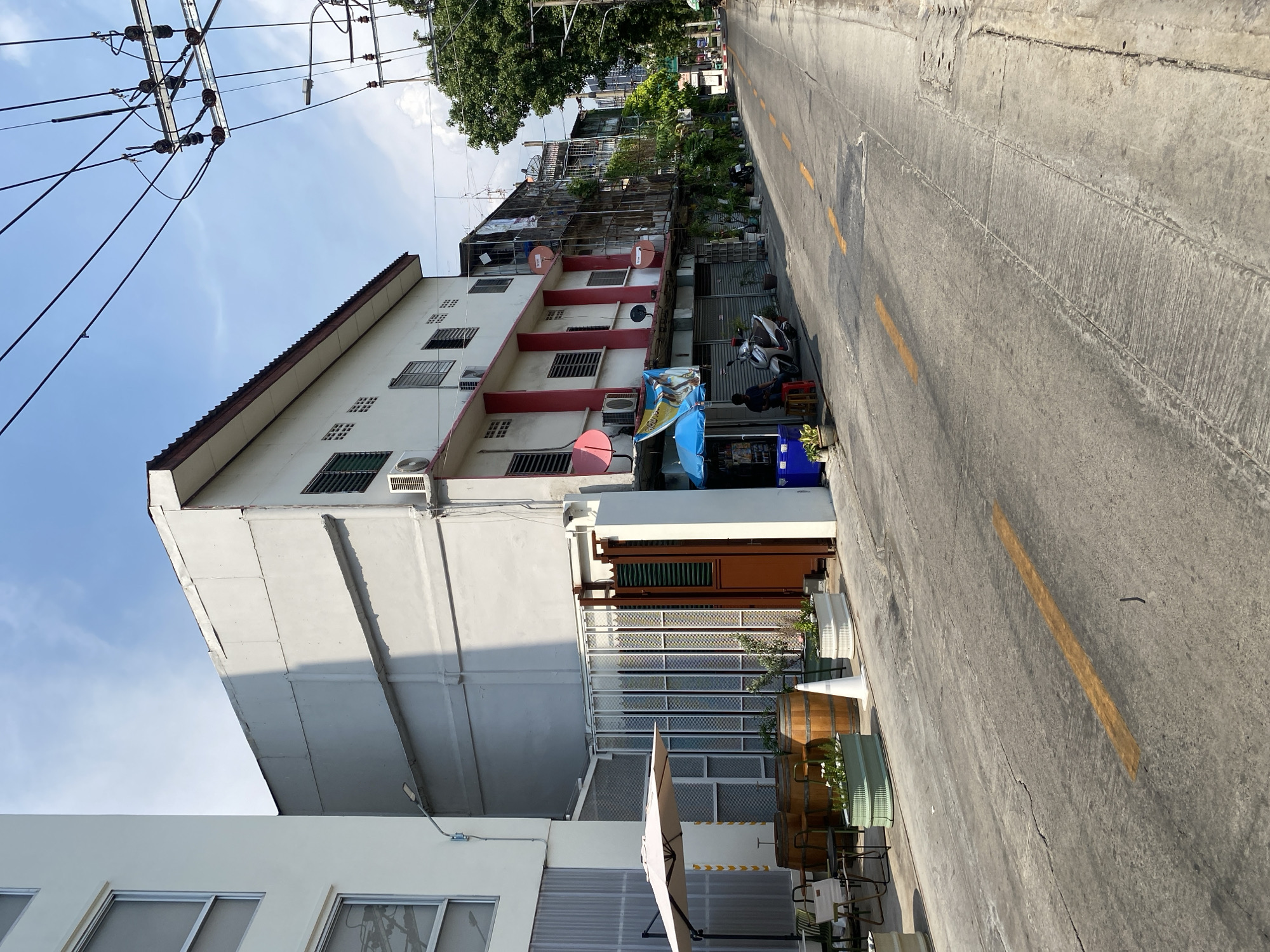 The image depicts a street scene in Bangkok, Thailand. A multi-story building with a white facade and red trim is visible on the left side of the frame. The building is equipped with multiple air conditioning units and satellite dishes. A gray concrete road with yellow lane markings occupies the right side of the frame. Electrical wires are visible overhead. A motorcycle is parked near a storefront. An umbrella and outdoor seating are in the foreground. The sky is partly cloudy.