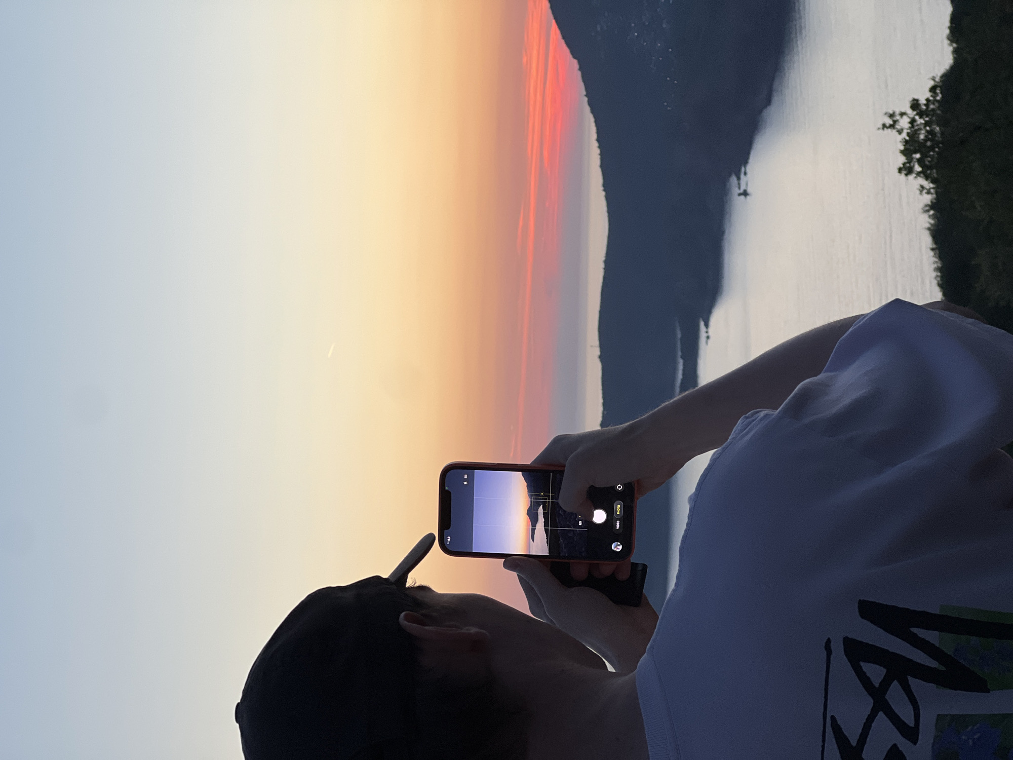 A person wearing a black cap and a white t-shirt is holding a red mobile phone, taking a picture of a scenic view in Šušanj, Montenegro. The view consists of a body of water reflecting the sky, a mountain range, and a sky displaying a gradient of colors, from light blue to orange and red, indicating either sunrise or sunset. The person is positioned on an elevated area, possibly a hill or a viewpoint. Vegetation is visible on the right side of the frame. The phone's screen displays a version of the landscape visible in the background.