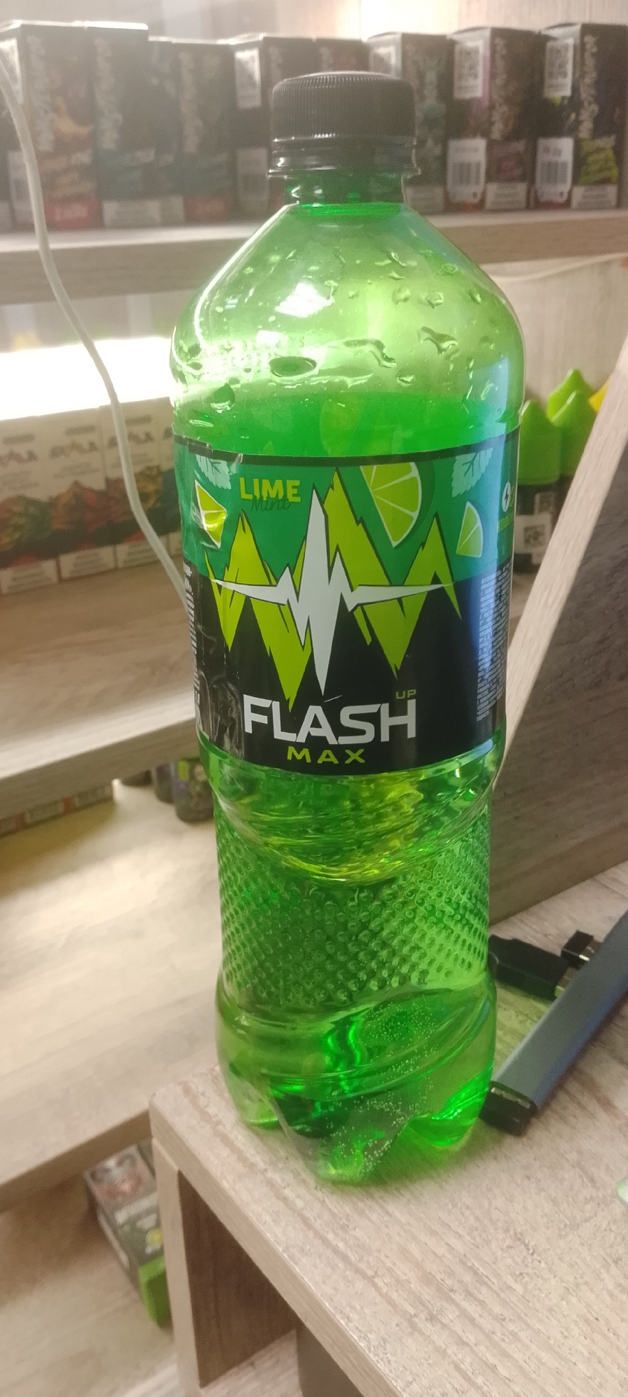 The image shows a close-up of a green plastic bottle of "FLASH UP MAX" lime-flavored energy drink. The bottle is condensation-covered and sits on a light-colored wooden surface. In the background, shelves are visible with various products, appearing to be in a retail setting. The overall lighting suggests an indoor environment, possibly a store or convenience shop. The location is identified as Krasnoyarsk, Russia. No people or actions are depicted.