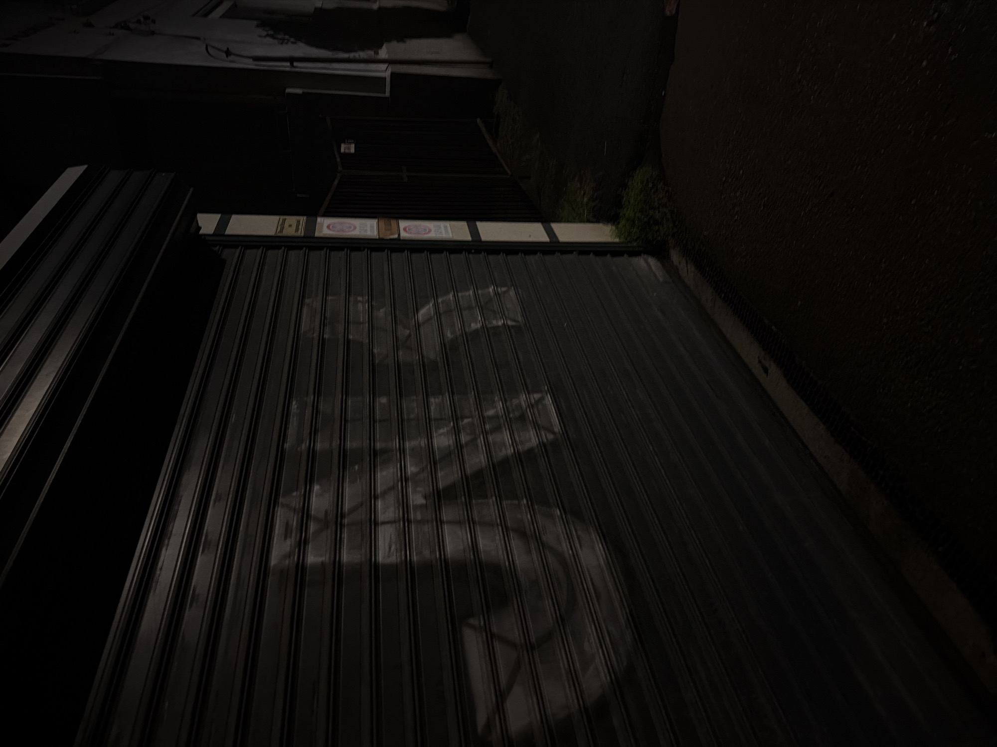 This media file captures a dark, static exterior scene at night in Pirgovo, Bulgaria. The primary subject is a large, dark corrugated metal roll-up door, likely belonging to a closed shop or garage.
Above the roll-up door, a horizontal panel features several signs. Prominently visible are at least two circular red and white warning or prohibitory signs, one distinctly showing a red circle with a diagonal line, suggesting a "No Entry" or similar restriction. Another similar sign has an indiscernible symbol within. Between these circular signs, a rectangular, lighter-colored sign with dark, illegible text is present, possibly indicating a business name or operating hours.
To the right of the building facade, a dark, textured wall or ground surface extends into the frame. Patches of green vegetation, possibly grass or weeds, are visible along the base of this wall. No people are present, and no activity is taking place. The scene is illuminated by very faint ambient light, which casts subtle reflections or patterns on the corrugated surface of the door. The overall impression is one of quietude and desertion, characteristic of a closed business area at night.