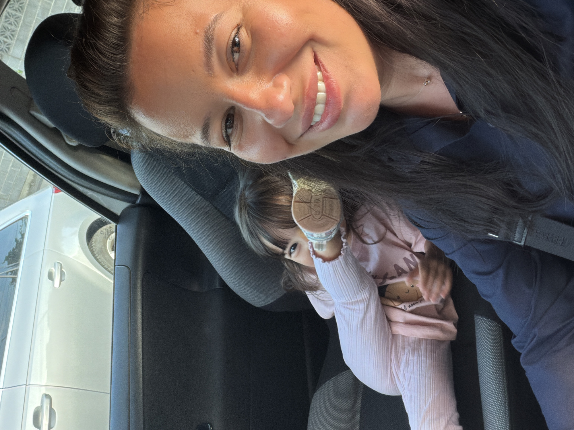 A selfie taken inside a car shows a woman in the foreground smiling at the camera, her right side is illuminated by sunlight. Behind her, a young child is seated in the back, their legs extended and partially obscuring their face. The child is wearing a pink outfit with a teddy bear graphic. The car's interior is visible, including black and grey seats and the doors. Outside the car window, another silver car is parked. The scene appears to be a roadside environment within the city of Burgas, Bulgaria.