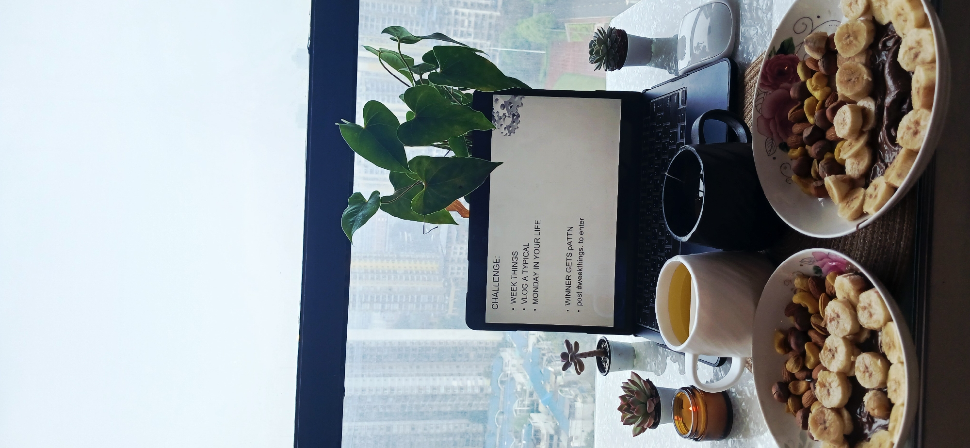 A desk in Jiulongpo District, China, features a laptop displaying a challenge, two mugs, two bowls of bananas and nuts, potted plants, a candle, and a computer mouse. The view outside the window shows a densely populated urban landscape.