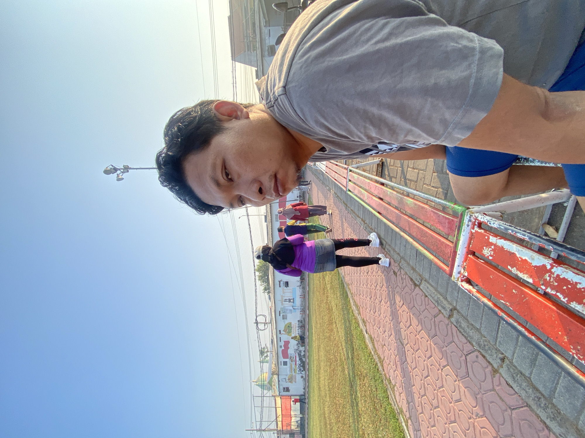A man sits on a red and white striped bench in what appears to be an outdoor sports or recreation area. He is wearing a grey t-shirt and blue shorts. In the background, a paved walkway is visible, with a grassy field to the left. Several people are walking along the path, including a woman in a purple top and denim skirt, and another in a red headscarf. A mural depicting various scenes is painted on a white wall behind them. The sky is clear and blue, suggesting it is daytime. The location is identified as Sumorame, Indonesia.