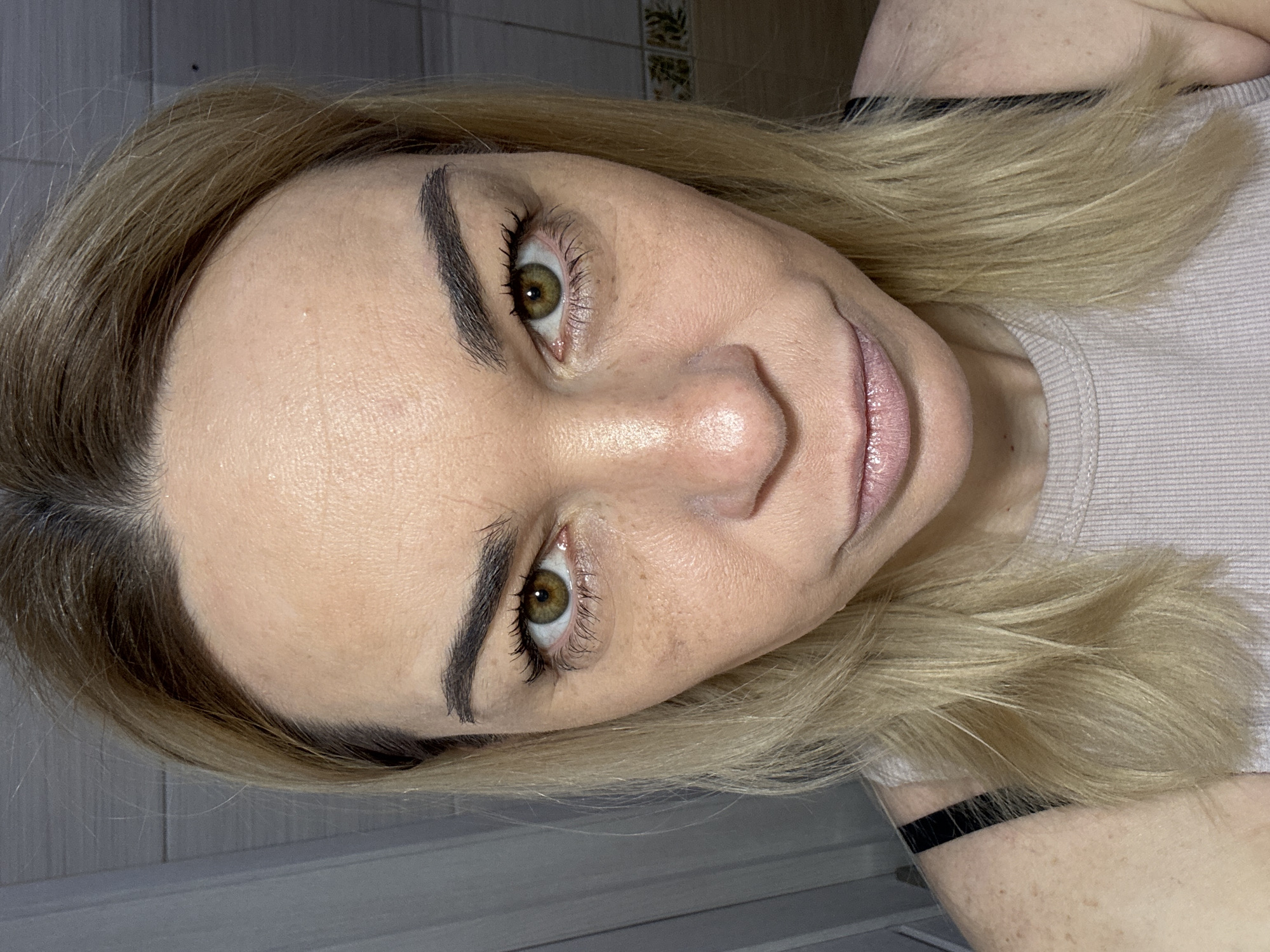 A close-up, angled view depicts a woman with light blonde hair and fair skin. Her eyes are hazel, and her eyebrows are darkly defined. She is wearing a light-colored, ribbed top with a black strap visible near her shoulder. The background is nondescript, featuring a grey surface in the lower left and a tile pattern near the top. There are no visible indicators to confirm the location is Vsevolozhsk, Russia.