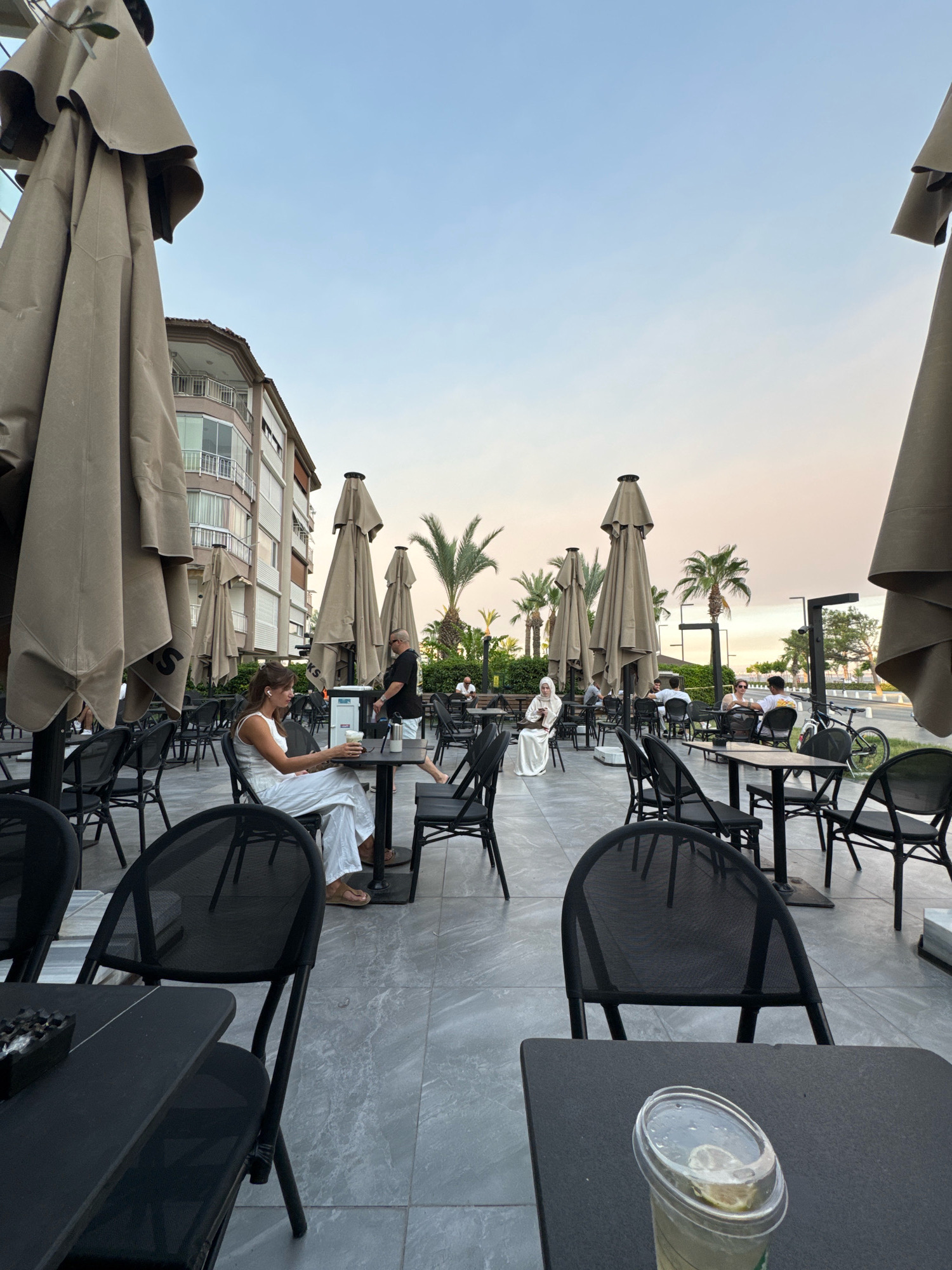 A cafe seating area is depicted on a grey tiled patio in Konyaaltı, Turkey. Large, tan umbrellas are set up to provide shade over the tables and chairs. Several people are seated at various tables, some interacting and others appearing to be alone.
In the foreground, a woman in a white outfit is seated at a table, holding a drink and looking to her left. To her right, a man in a black t-shirt and shorts stands near another table, talking to someone. Further into the background, a woman in a white hijab is seated at a table by herself. Other patrons are visible at tables in the mid-ground and background.
The environment includes palm trees and a building with balconies and windows in the mid-ground. In the background, there is a road with a bicycle parked nearby, and a glimpse of the sky suggests it is late afternoon or early evening, with a soft gradient of blue and pink. The cafe appears to be an outdoor section of a Starbucks, indicated by the branding on the umbrellas.