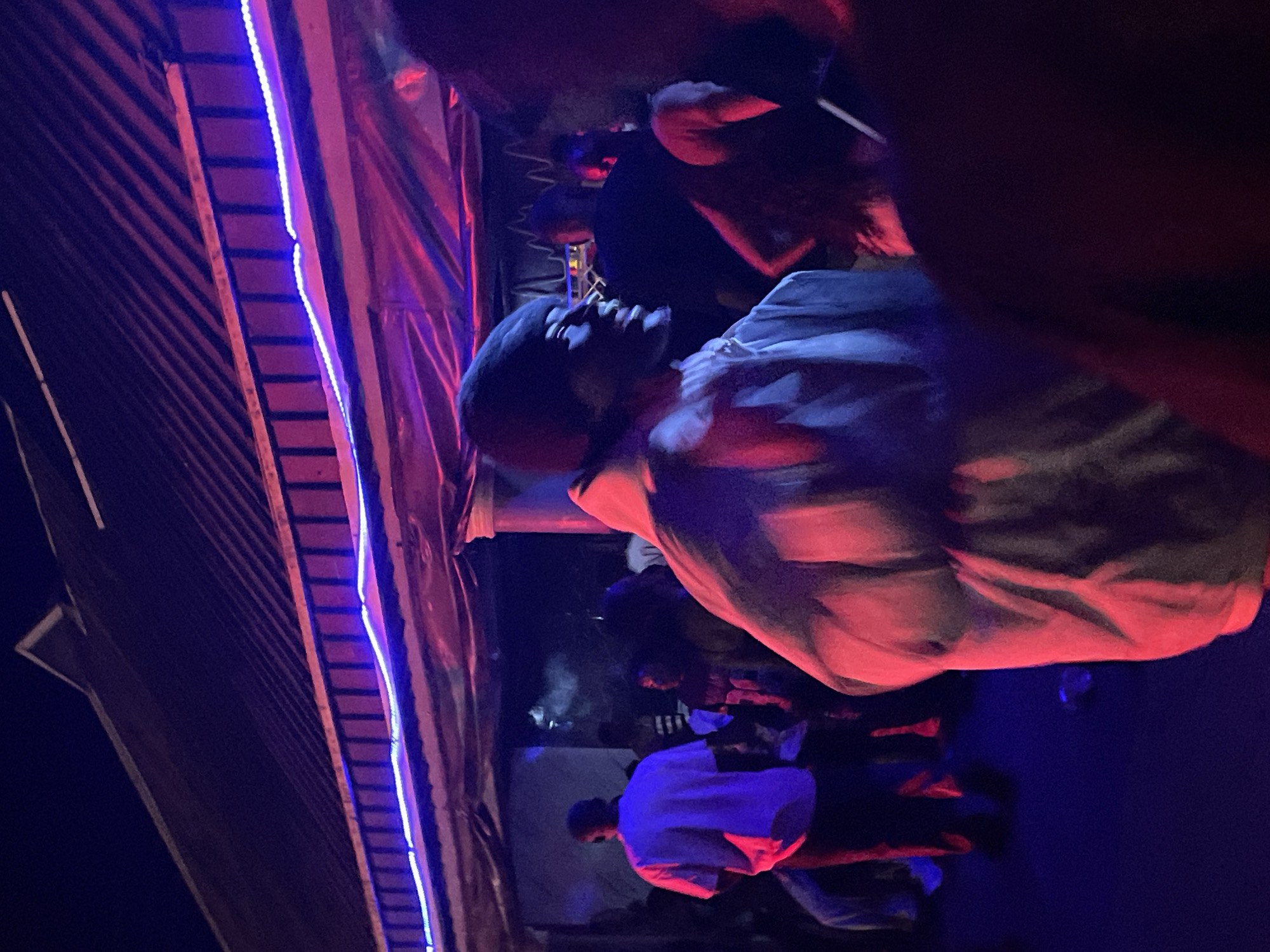 The image depicts a nighttime scene at an outdoor event in Durumi, Nigeria, likely a gathering or party. The setting is illuminated by vibrant blue and pink neon lights strung along the roofline of a structure that appears to be a covered outdoor area, possibly a pavilion or a temporary stall. The ground is dark, suggesting it is nighttime, and there are indications of a gathering of people, though their faces are mostly obscured or out of focus.
In the foreground, a person is seen from the shoulders up, facing to the right, with their profile illuminated by the colorful lights. They are wearing a dark beanie and a light-colored, possibly white or pale grey, puffy jacket or hoodie. Their expression is animated, with their mouth open as if speaking or reacting to something.
Further back, to the left, a person in a white short-sleeved shirt and dark pants is standing, facing away from the camera. To their right, a person with curly hair is partially visible, wearing a dark top with some markings. Other figures are indistinct in the background, suggesting a crowd. The overall atmosphere appears lively and social, characteristic of an evening event or celebration.