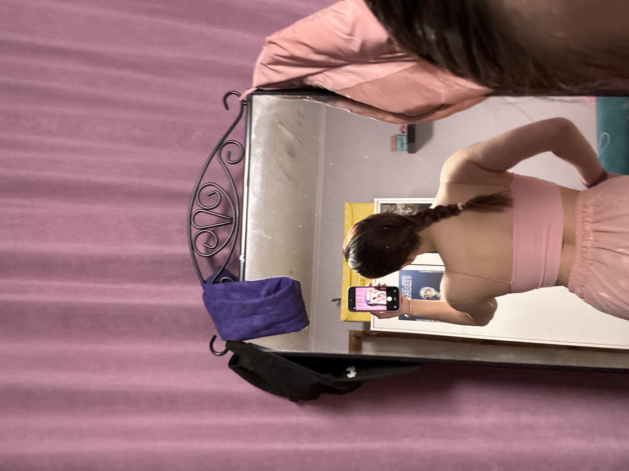 A person is taking a picture in a mirror. The person is wearing a pink top and bottom set. The person has a ponytail. The mirror is hanging on a wall with a pink sheet or blanket. There is a purple cloth item hanging on the mirror's frame, along with a black object. In the mirror, there is a framed document on the wall. The person is taking a picture of themselves in the mirror with their cell phone. The photo was taken in Прикамский, Russia.