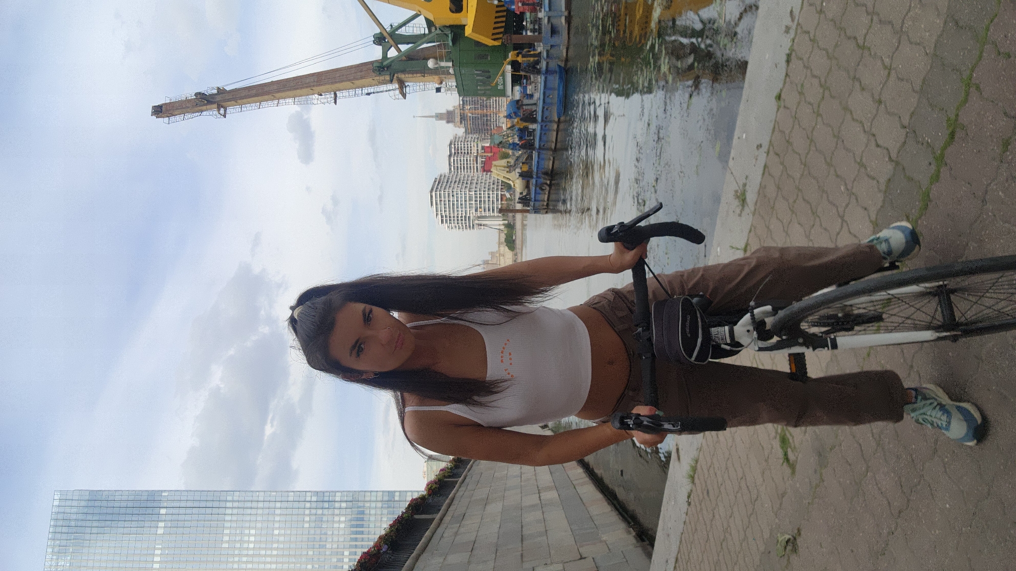 In the image, a young woman with long dark hair is sitting on a bicycle next to a body of water. She is wearing a white crop top and brown pants. The bicycle is black with white accents. Behind her, there is a large crane and several buildings in the background. The water appears calm, and the sky is overcast. The ground is paved with a textured pattern. The woman is looking towards the camera with a neutral expression. There are no visible signs or text in the image. The overall impression is that of a casual day out, possibly in an urban or industrial waterfront area. The lighting suggests it might be daytime, but the cloudy sky makes it difficult to determine the exact time.