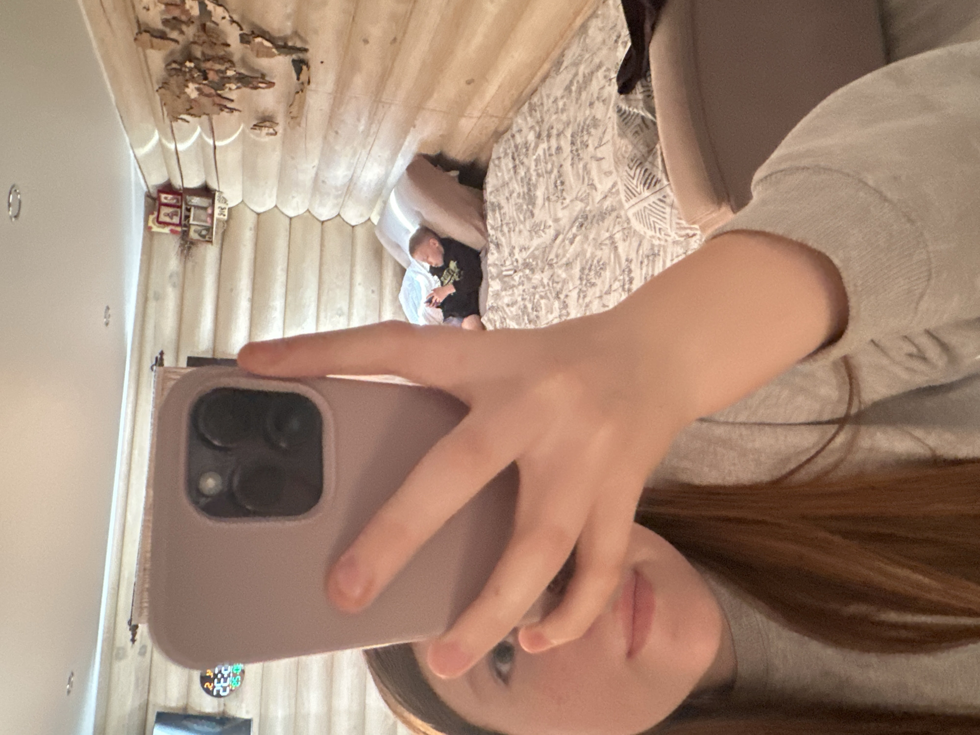 A person with long brown hair and wearing a grey sweatshirt is taking a selfie using a purple iPhone. A child wearing a black shirt is lying on a bed with a floral pattern bedspread, and is holding a cell phone. The room has wood-paneled walls and ceiling. Hanging from the ceiling are decorations including small frames and a wooden cutout. A television is visible in the lower left. The location is likely in Ольховка, Russia.