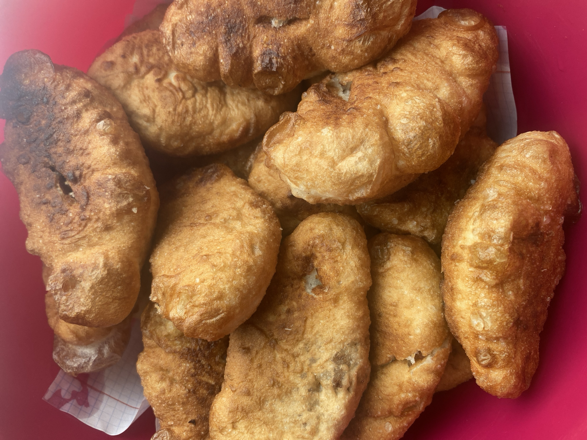 A close-up, overhead shot shows a pile of fried dough pastries, likely belyashi, in a bright pink bowl. The pastries are irregular in shape and golden brown with some darker, more caramelized spots. They appear to be soft and slightly greasy. A piece of white checkered paper is visible under some of the pastries on the left side of the frame. The background is a solid, vibrant pink. The image does not contain any information about the city of Бетюнцы, Russia, or any people or actions.