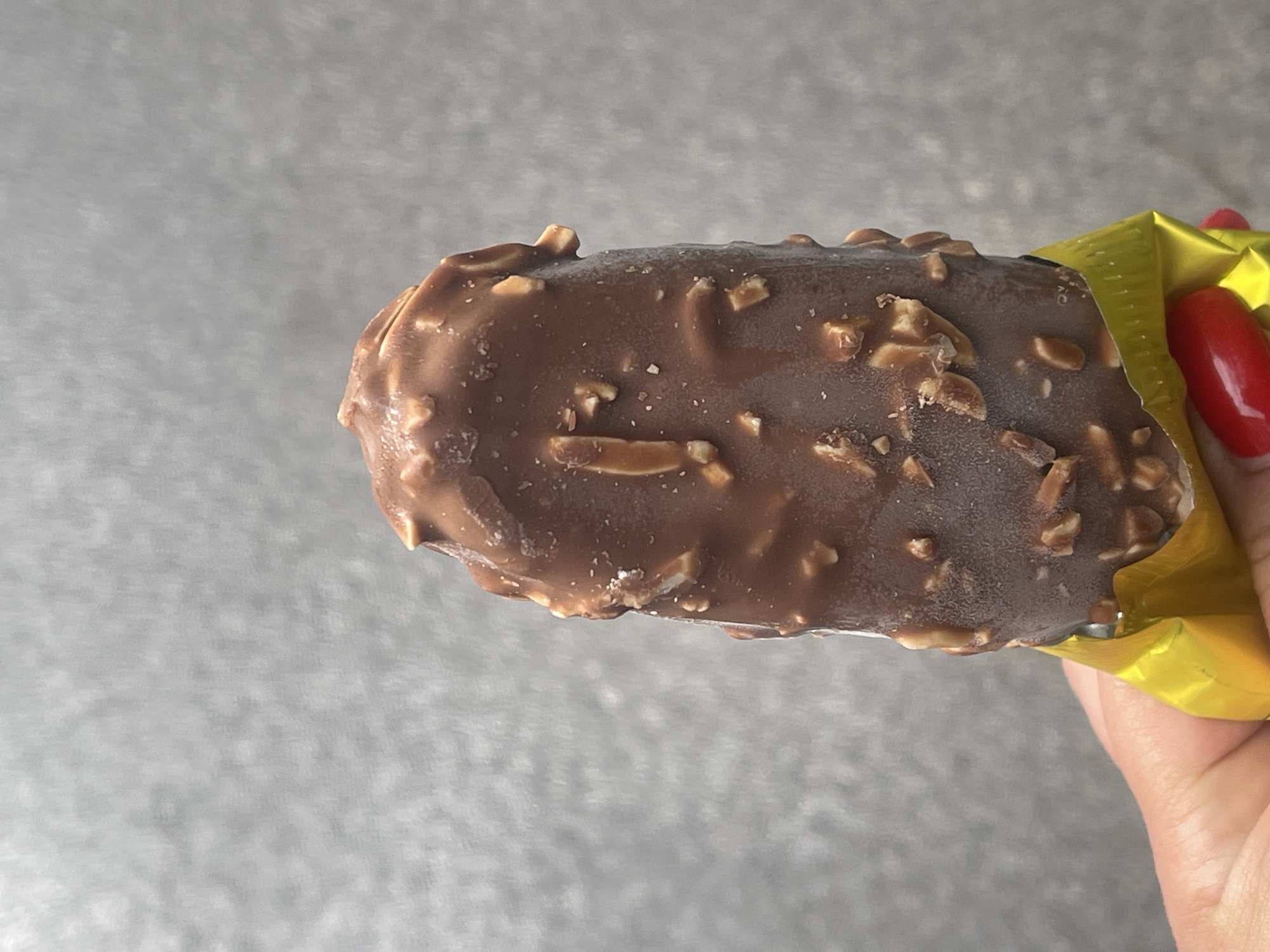 The image presents a close-up view of an ice cream bar held by a person's hand against a plain, light gray background. The primary subject is a rectangular ice cream bar, coated in milk chocolate and generously covered with visible chopped nuts, likely almonds or peanuts. The chocolate coating shows slight condensation or frost, indicating the dessert's cold temperature. The ice cream bar is partially unwrapped from its packaging, which is a bright, crinkled golden-yellow foil. A hand, distinguished by fingers with prominent bright red polished fingernails, is visible on the right side of the frame, holding the ice cream and its wrapper.
The activity depicted is the holding of the ice cream bar, suggesting it is either being presented for consumption or has just been unwrapped. There are no other visible subjects or activities. The background is uniformly light gray with a subtle, soft texture, keeping the focus entirely on the ice cream and the hand. No specific location cues are visible within the frame, beyond the contextual information that the image originates from Poltava, Ukraine. There is no visible text on the product or its packaging. The lighting appears even, highlighting the details of the ice cream and the hand.