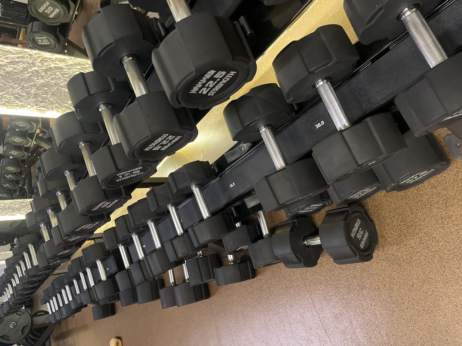 The image captures a well-organized dumbbell rack within a gym or fitness center, likely located in North Kuta, Indonesia. Rows of black, hexagonal dumbbells with silver handles are neatly arranged on multiple tiered racks, receding into the background on both the left and right sides of the frame. The dumbbells vary in weight, indicating a comprehensive set for strength training.
The floor is a light brown, textured surface, typical of gym flooring. The background features strong reflections, suggesting large mirrors on the walls or ceiling, which also reflect overhead artificial lighting, contributing to a bright indoor environment. There are no people visible, implying the gym is either empty or the focus is solely on the equipment.
Visible text on the dumbbells clearly indicates the brand "HAMMER STRENGTH" and specific weights. Prominently displayed weights include "22.5 STRENGTH", "30.0", "32.5", and "35 STRENGTH". Some other text fragments, possibly distorted reflections or partial views, include "OS" and "2.51". The overall impression is one of orderliness and readiness for exercise.