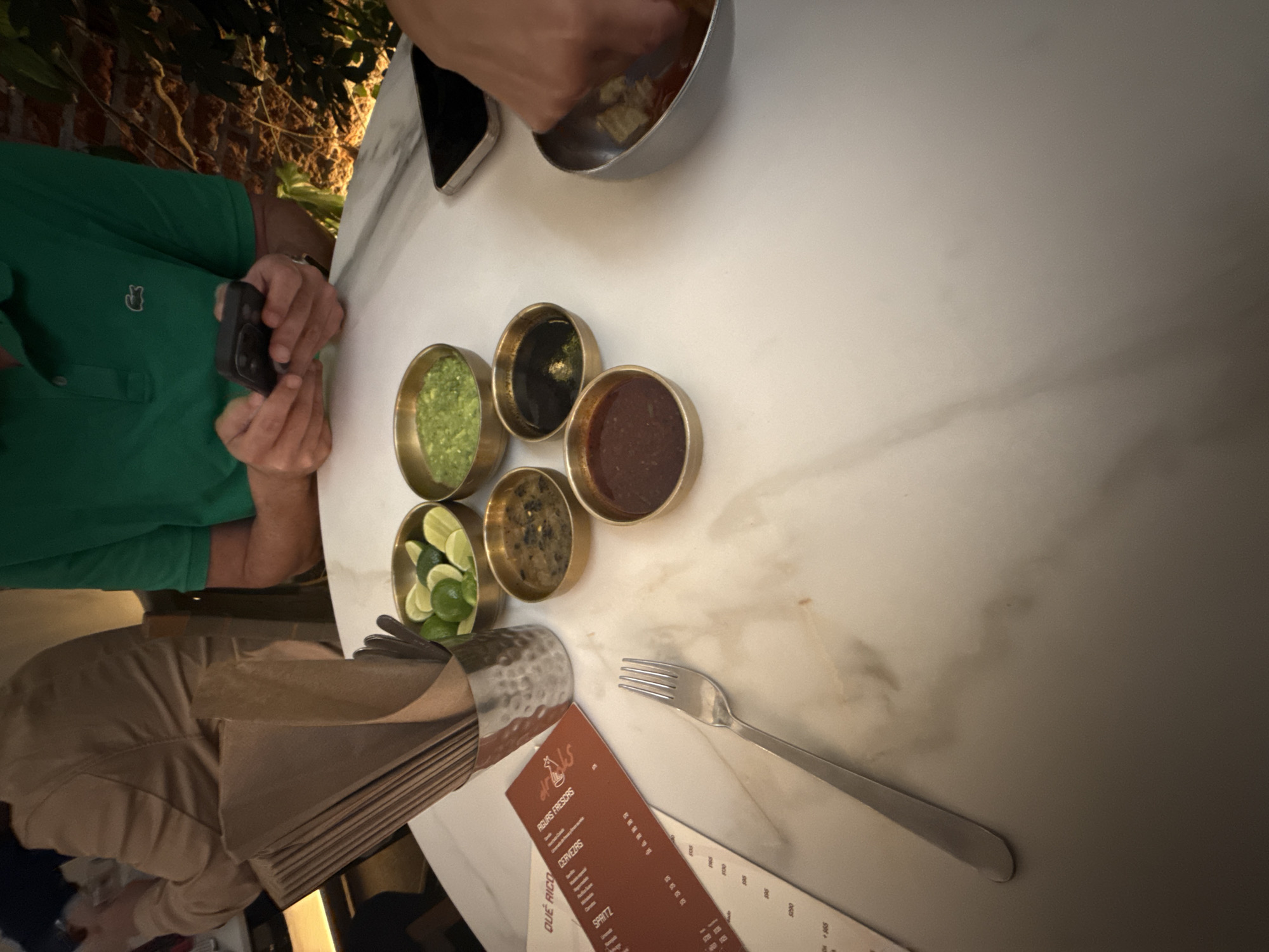 A table in a restaurant in Mexico City, Mexico, contains a menu, a fork, a napkin holder with napkins, five small bowls of different sauces or condiments (one with lime slices), a bowl with food being eaten, and two phones. Two people are at the table. One person is wearing a green polo shirt and holding a black phone. The other person is only visible by their hand reaching into a bowl of food. The restaurant appears to have plant life nearby.