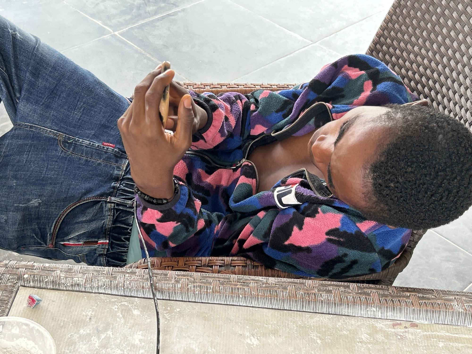 A person is lying on a woven chair, looking at a phone. They are wearing blue jeans and a colorful, patterned fleece jacket. The background shows a tiled floor, suggesting an outdoor or semi-outdoor setting, possibly in Ihiagwa, Nigeria.