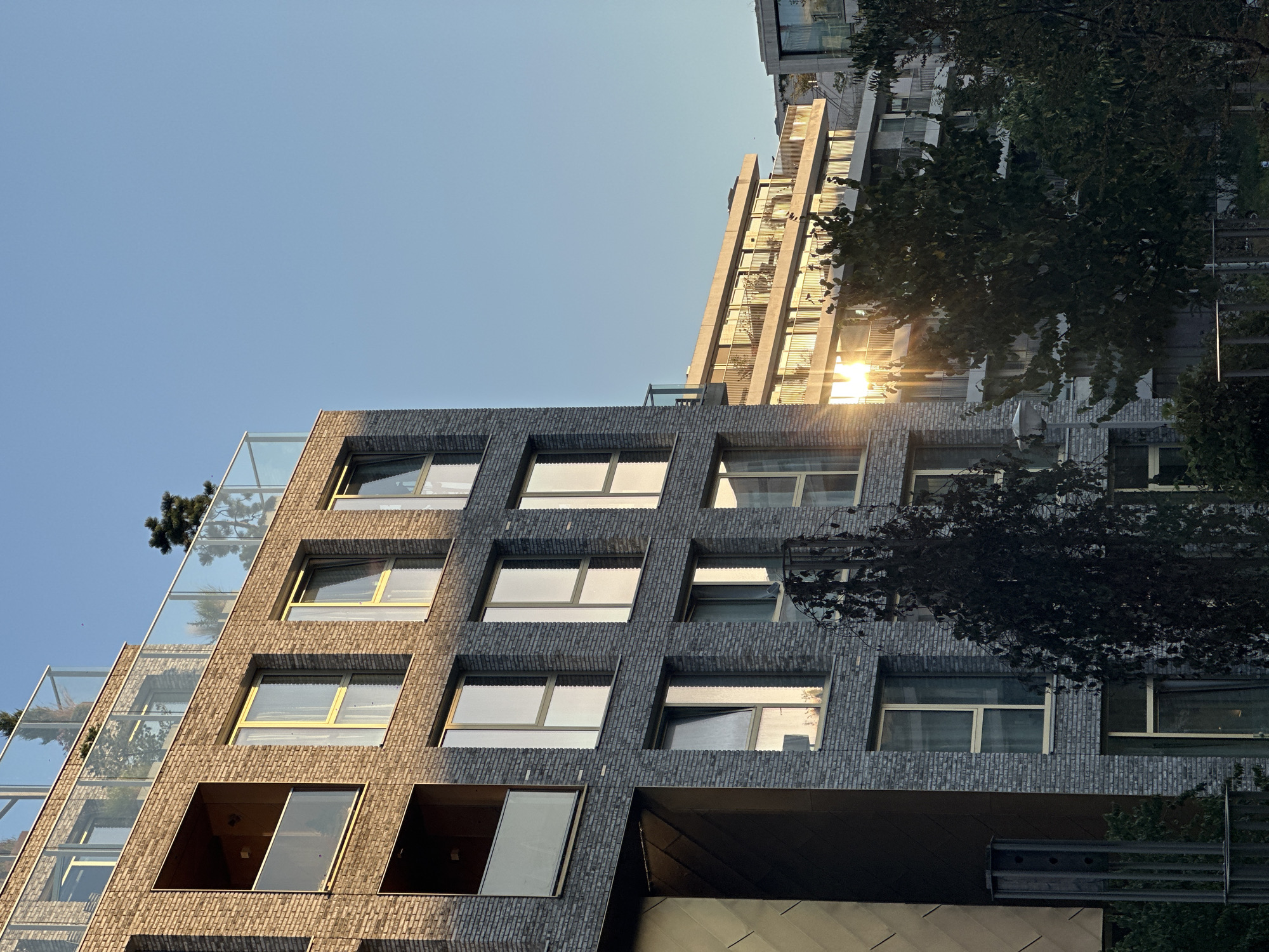 The image depicts a close-up view of a modern brick building with multiple floors and balconies, illuminated by the warm glow of the setting sun. The building's facade is constructed of grey bricks, and it features numerous large windows, some with yellow frames. Several balconies with glass railings are visible, with some containing potted plants and trees. The sky is a clear, pale blue.
In the background, another building with a lighter facade and similar balcony structures is visible, also catching the sunlight. Trees with dark green foliage are in the foreground and middle ground, partially obscuring the lower levels of the buildings. The overall scene suggests an urban residential area, likely in the city of Amsterdam, Netherlands, given the architectural style. There are no people visible in the image. The sunlight creates strong reflections on the windows, adding a dynamic element to the composition.