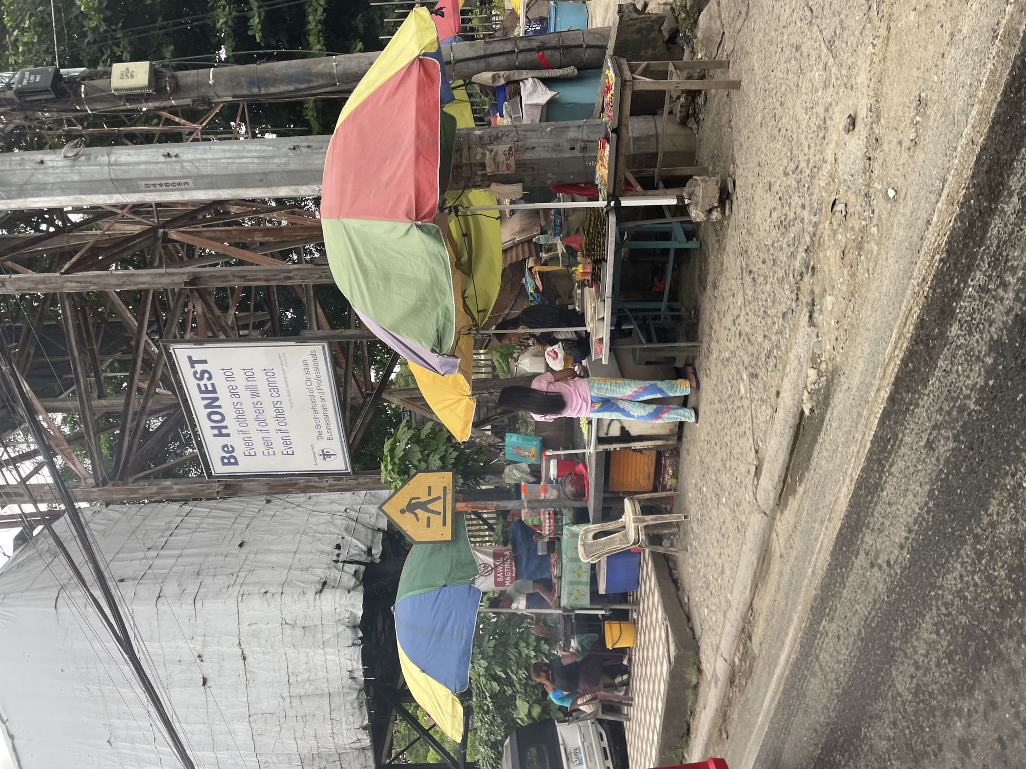 The image depicts a street scene in Davao City, Philippines, likely a roadside market. In the foreground, a paved road with some debris is visible. Behind it, a sidewalk area is occupied by several vendors selling food items under colorful umbrellas. One stall features food displayed on tables, with a vendor attending to customers. Another stall appears to be grilling food. Several people are present, some seated, and one person is standing and facing away from the camera. A white vehicle is parked on the left side of the image. In the background, a large white structure, possibly a building under construction or renovation, is covered with a tarp. Electrical poles and wires are prominent. A sign on a pole reads "Be HONEST Even if others are not Even if others will not Even if others cannot" and mentions "The Brotherhood of Christian Businessmen and Professionals". A pedestrian crossing sign is also visible. The overall impression is of a bustling commercial area on a cloudy day.