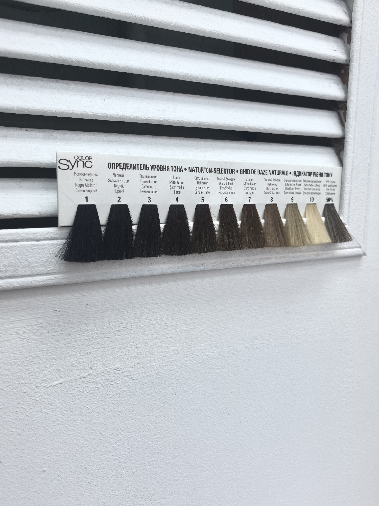 The image displays a hair color swatch chart from the "COLOR Sync" brand, presented in a salon or retail setting in the city of Chaikovsky, Russia. The chart, labeled "ОПРЕДЕЛИТЕЛЬ УРОВНЯ ТОНА" (Level Tone Indicator) in multiple languages, features ten distinct synthetic hair swatches, numbered 1 through 10, illustrating a gradient of hair colors from dark to light.
Swatch 1 is "Иссиня-черный" (Bluish-black), followed by Swatch 2, "Черный" (Black). The progression continues through various shades of brown: Swatch 3, "Темный шатен" (Dark Brown); Swatch 4, "Шатен" (Brown); and Swatch 5, "Светлый шатен" (Light Brown). The chart then transitions into blonde tones: Swatch 6, "Темный блондин" (Dark Blond); Swatch 7, "Блондин" (Blond); Swatch 8, "Светлый блондин" (Light Blond); Swatch 9, "Очень светлый блондин" (Very Light Blond); and Swatch 10, "Очень-очень светлый блондин" (Extra Light Blond). Two additional swatches to the right of number 10 are marked "50% Седины" (50% Grey), indicating a mix of dark and grey strands.
The white rectangular chart is positioned against a background primarily composed of horizontal white slats or louvers, likely part of a ventilation grille or blind, with shadowed gaps between them. Below the chart, and forming the lower portion of the frame, is a plain, textured white wall surface. No individuals or active interactions are visible in the scene. The lighting is bright and even.