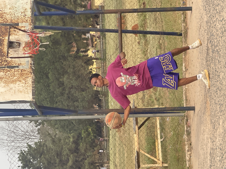 An outdoor basketball court in Umunekwu, Nigeria, features a single male subject, appearing late teens to early twenties, with dark skin and short dark hair. He wears a magenta t-shirt displaying a reddish-brown robot graphic, purple basketball shorts with "LAKERS" in gold lettering, and light-colored athletic footwear. He holds a worn, orange basketball in his left hand, with his arm extended. His head is tilted back, eyes squinted or closed, and mouth closed, suggesting concentration. He is captured in mid-action, balancing on his left foot with his right foot slightly lifted. Behind the subject, a heavily rusted basketball backboard with a red rim and a white, frayed net is visible, supported by blue poles. A chain-link fence defines the court boundary, behind which are a grassy field, numerous green trees, some bare deciduous trees, and faint human figures. A light-colored wooden bench-like structure is present to the left. The court surface is worn, light-colored asphalt or concrete.