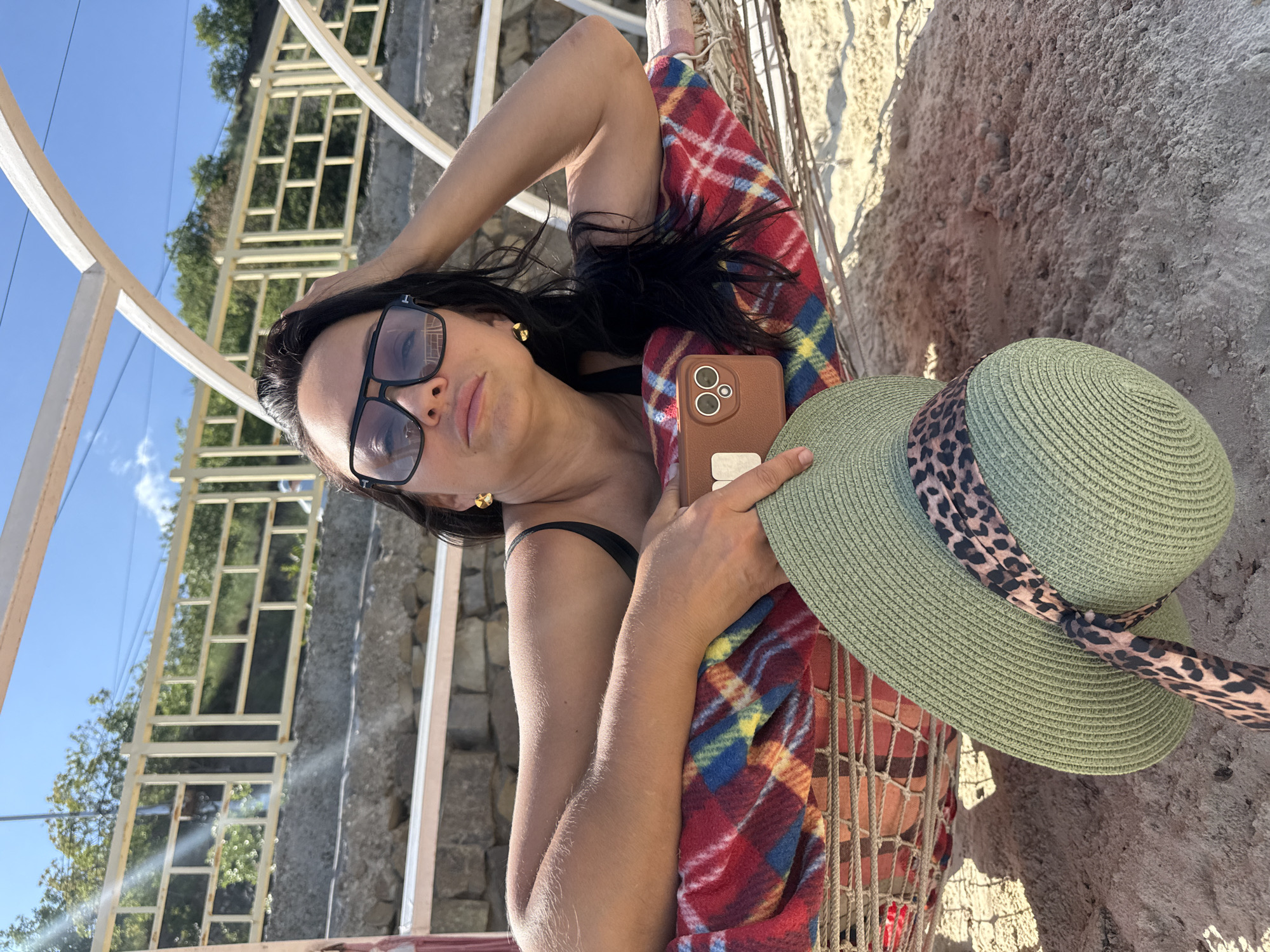 A young woman is depicted in a close-up shot, lying on her back, angled to her left, likely in a hammock indicated by the net-like structure beneath her. She wears large, dark square sunglasses and a black bikini top. Her long, dark hair is styled away from her face, and a gold stud earring is visible on her left ear. She is partially covered by a red, blue, and yellow plaid blanket or towel.
In her right hand, she holds a brown smartphone with multiple visible camera lenses, while her left hand supports a light green wide-brimmed straw hat adorned with a leopard print band. She looks towards the camera with a relaxed or contemplative expression.
The background features a light-colored railing or pergola structure, with green trees and a clear blue sky visible beyond it. To her right and behind the hammock, there is a rough, light grey, textured wall or cliff face, possibly made of concrete or rock, with sandy or rocky ground below. The scene is brightly lit by natural sunlight, indicating a clear, sunny day outdoors. No visible text is present in the image.