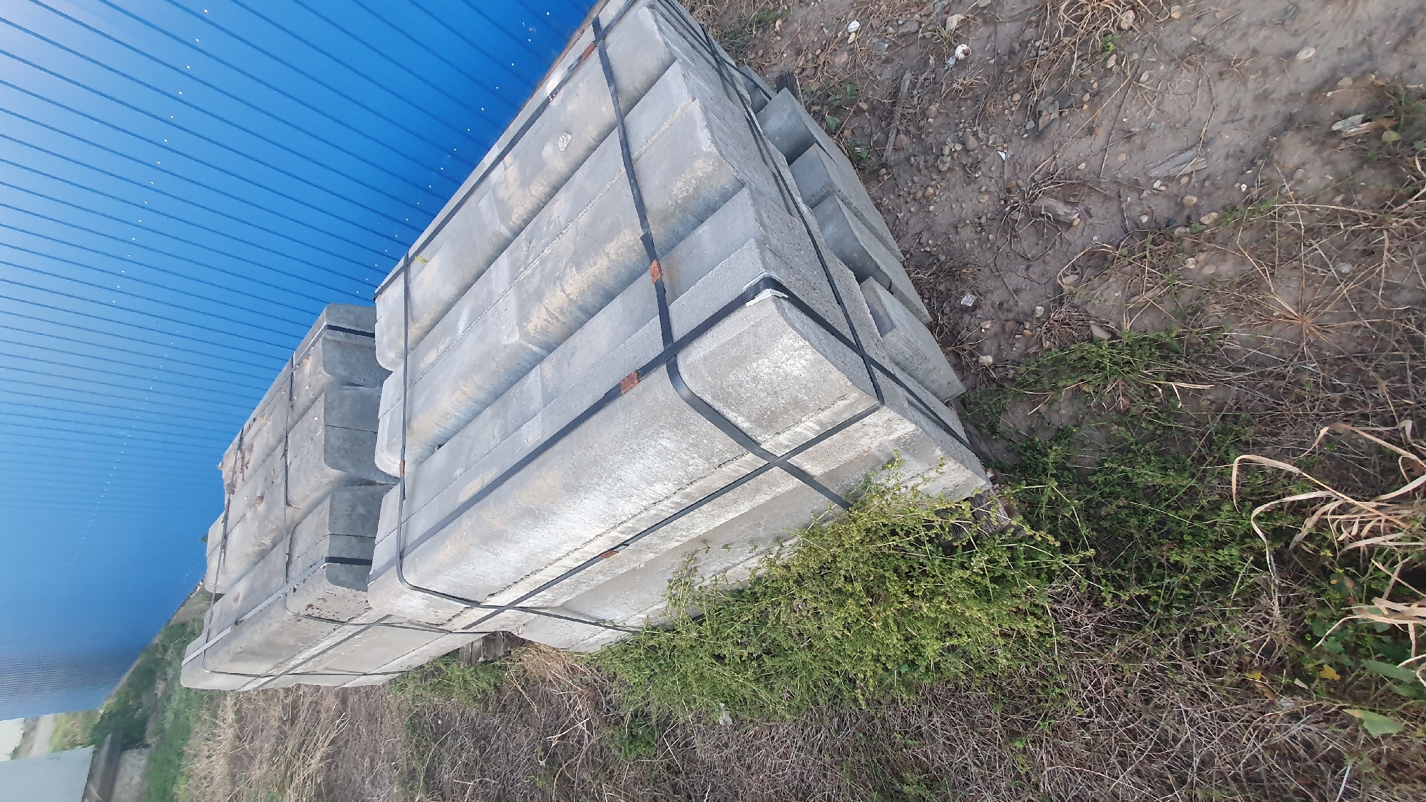 The image depicts a stack of concrete curbs placed outdoors, with a blue corrugated metal wall in the background. The curbs are gray, rectangular with a curved top edge, and are banded together with black plastic straps. They are stacked in two main piles, with one pile partially obscured by green vegetation. The ground around the curbs is covered with dry grass, dirt, and small stones. The overall scene suggests an outdoor storage area, possibly at a construction site or material yard.