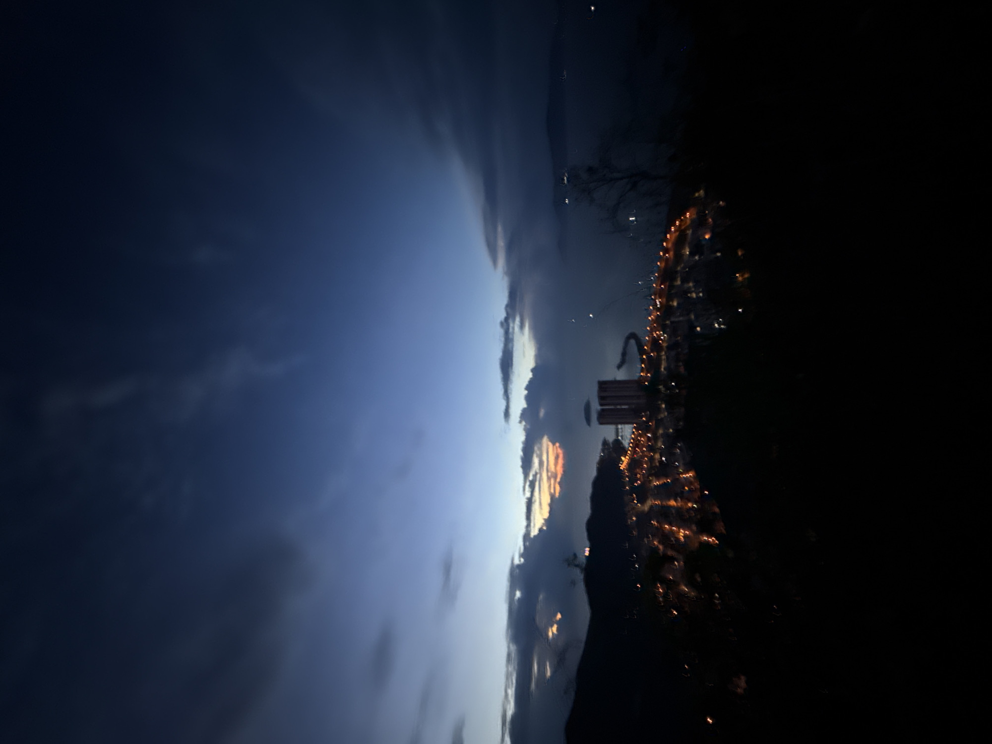 The image shows a nighttime view of Bac Nha Trang Ward, Vietnam. The sky transitions from a dark blue-grey to a lighter blue with clouds near the horizon where the sun has set. Building lights are visible on the right side of the image, creating a blurred effect due to the camera's movement. Some tall structures are distinguishable against the horizon. The bottom portion of the image is obscured by darkness.